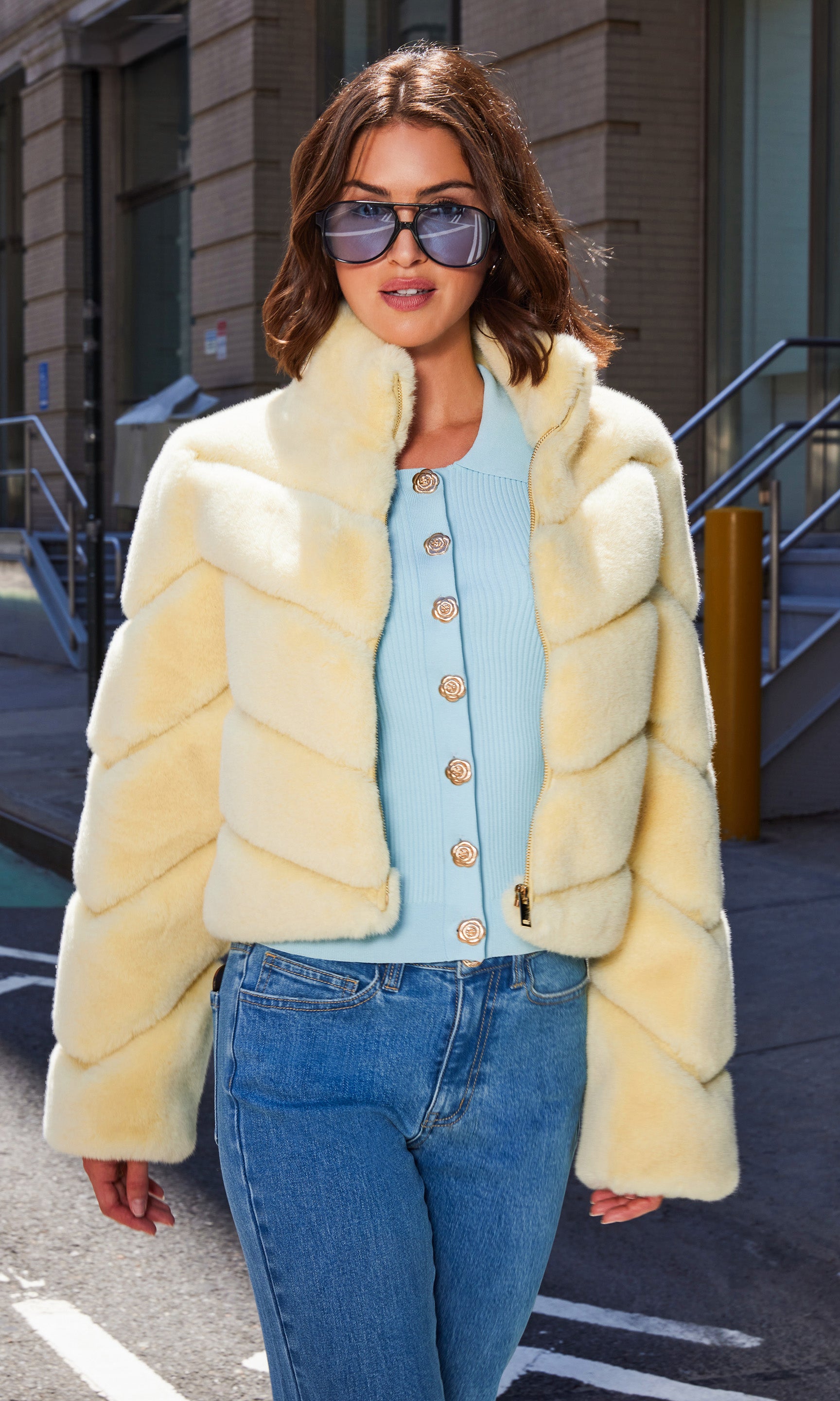 The model stands in a front view wearing the Brianna Faux Fur Jacket in pale yellow layered over a pastel blue cardigan and slim blue jeans, hands tucked into pockets for a confident stance.