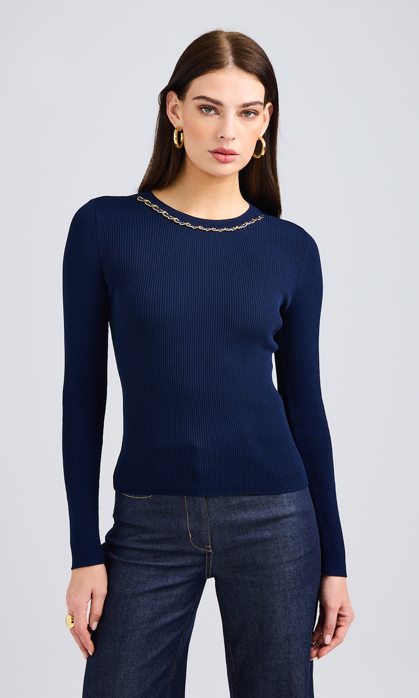 Woman wearing a ribbed sweater with long-sleeves and a chain at the neckline paired with jeans.