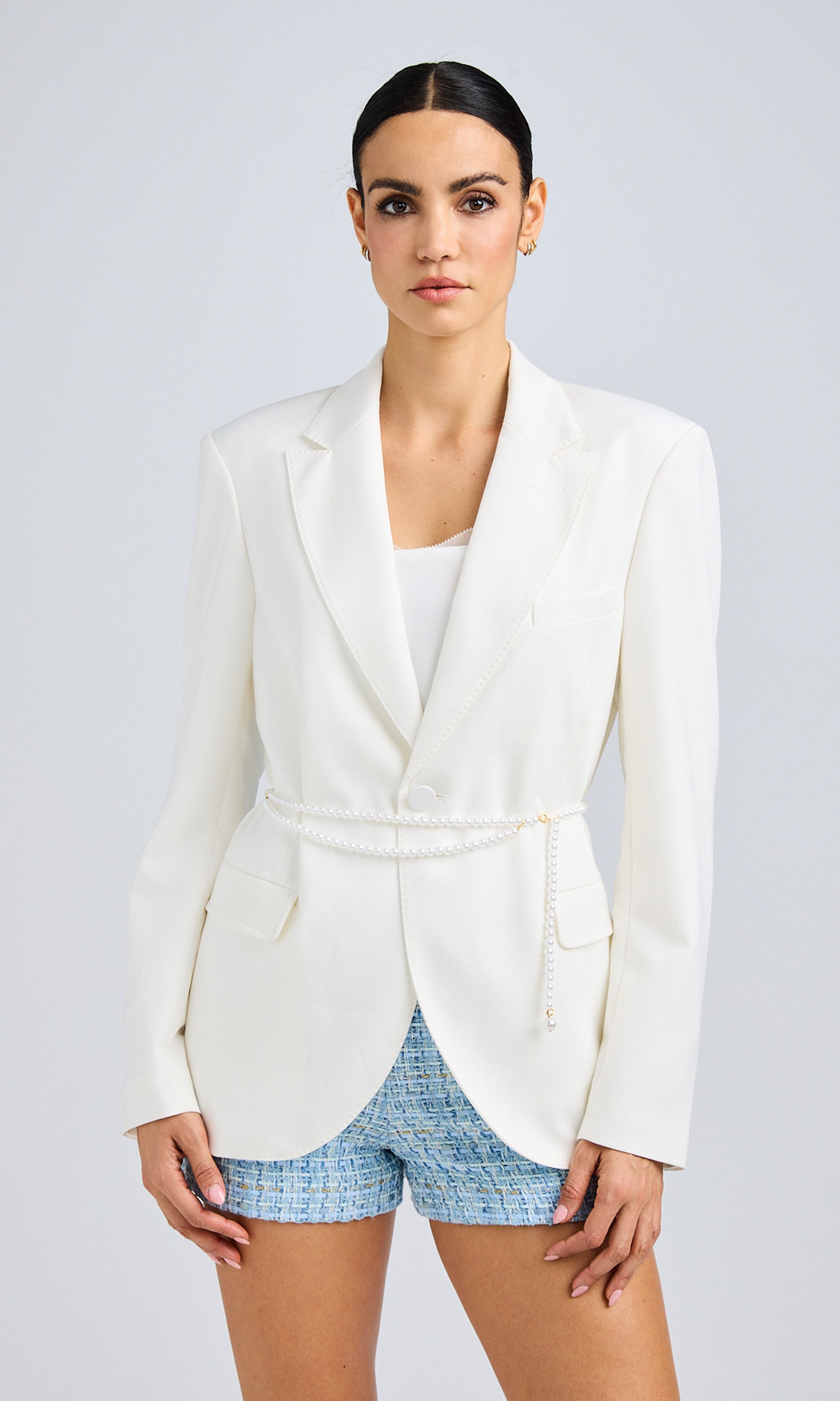 Front view of a model wearing a white belted blazer with blue textured shorts against a plain background.