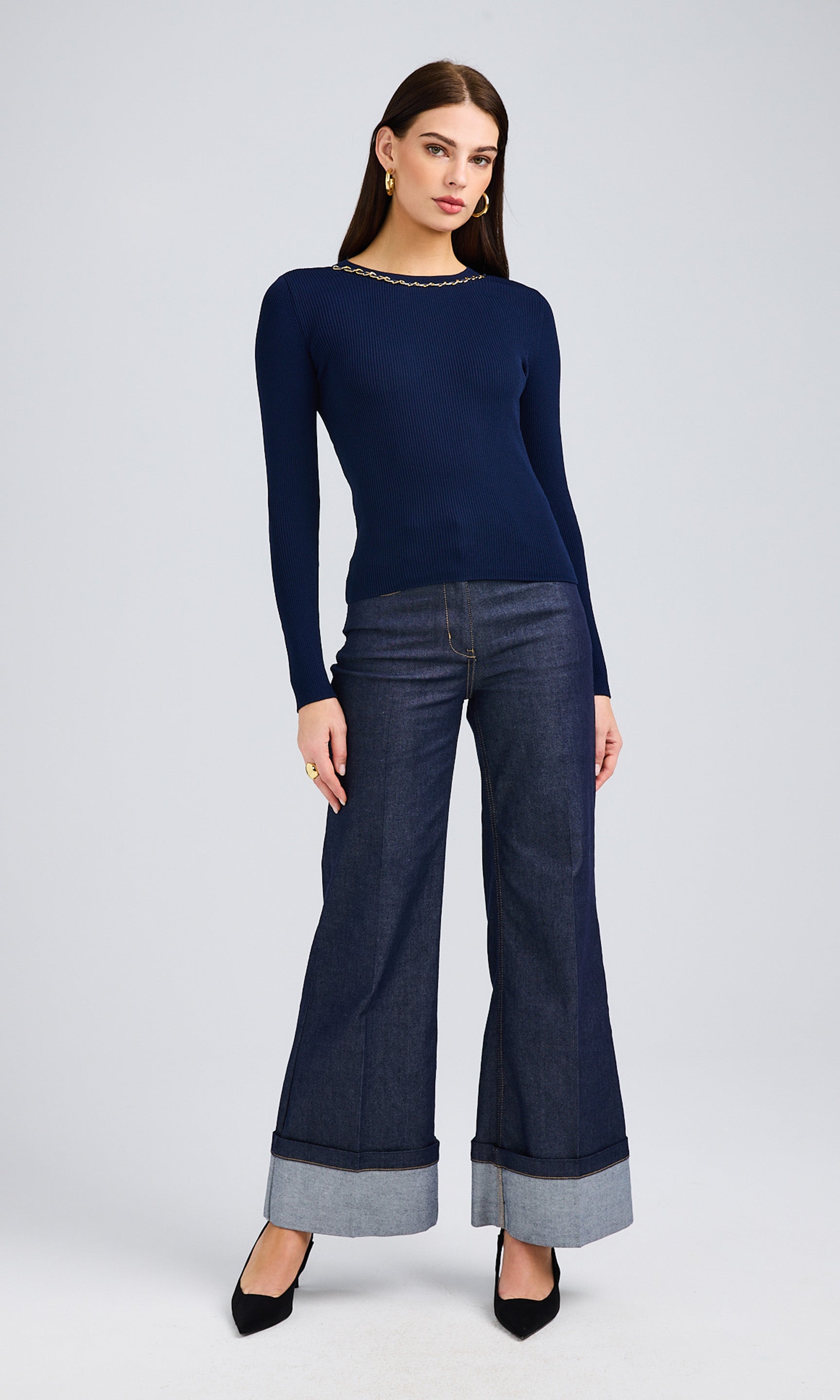 Dark-haired woman wearing a navy sweater with gold chain details and wide-leg cuffed jeans.