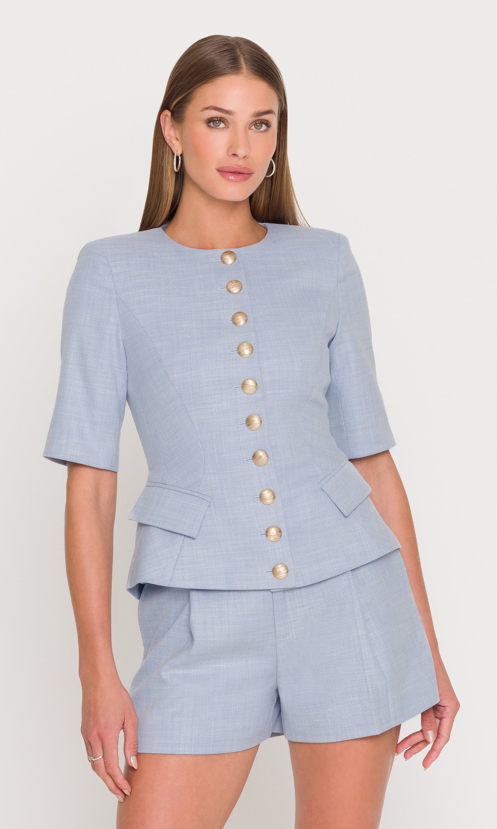 Model wears the Adriana Chambray Suiting Jacket in chambray blue in a front view, showing the short sleeves, peplum waist, and gold button trim.