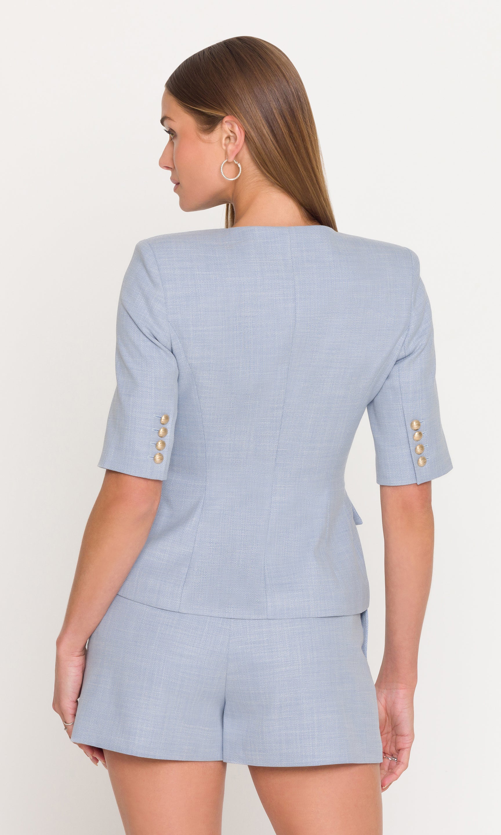 Model wears the Adriana Chambray Suiting Jacket in chambray blue in a back view, showing the clean tailored lines and short sleeve shape.