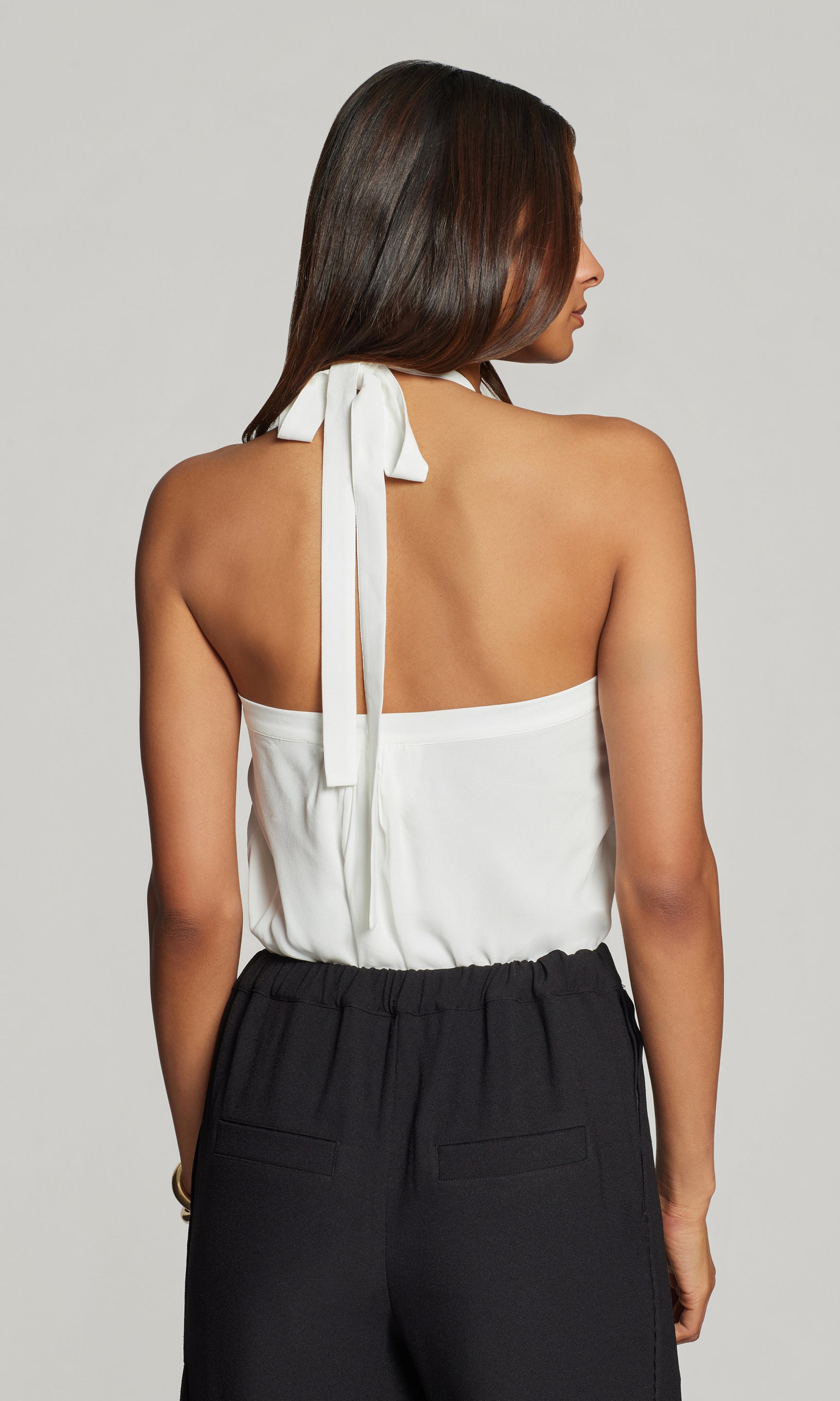 Back view of woman wearing a white halter top with black pants on a plain background