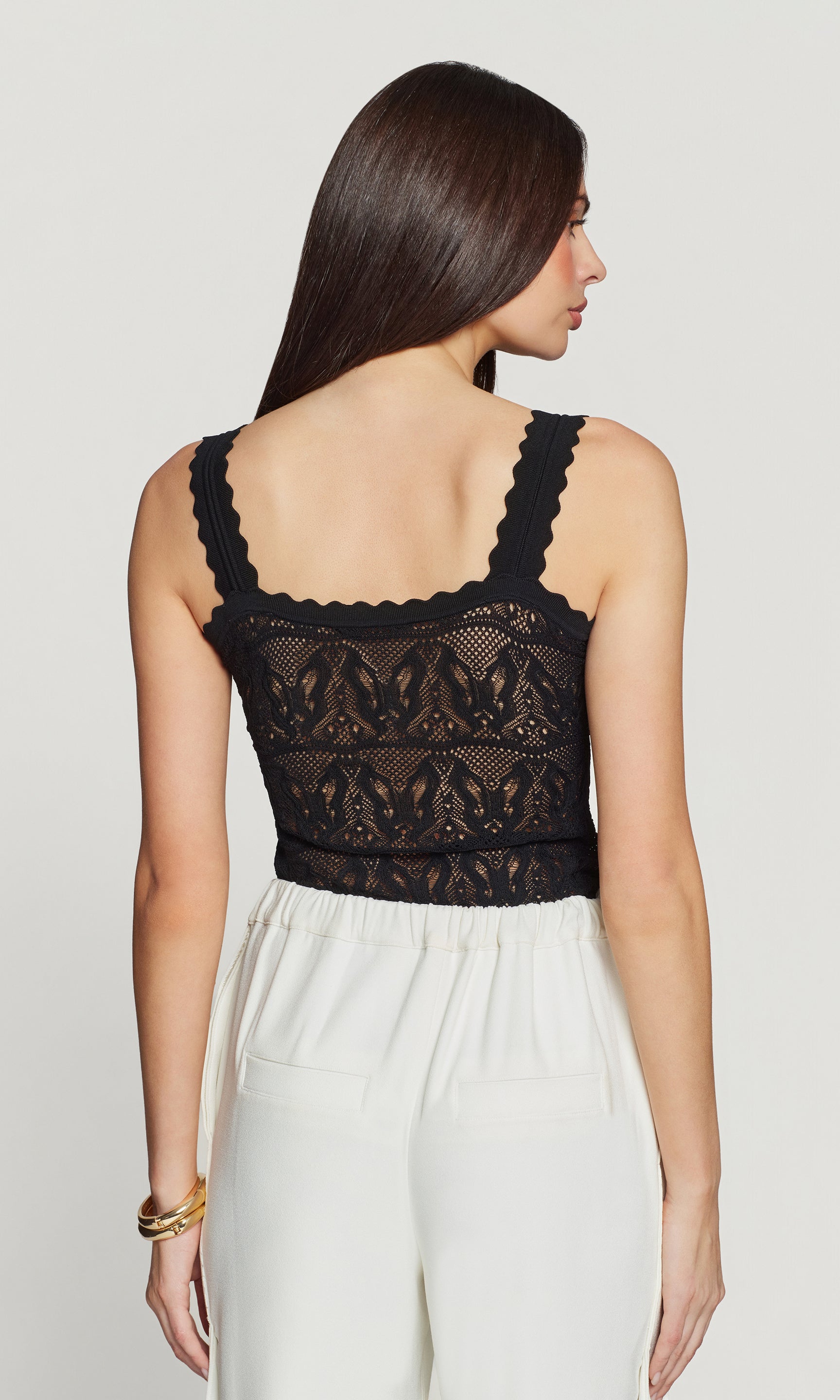 Back view of woman wearing a black lace top and white pants on a plain background