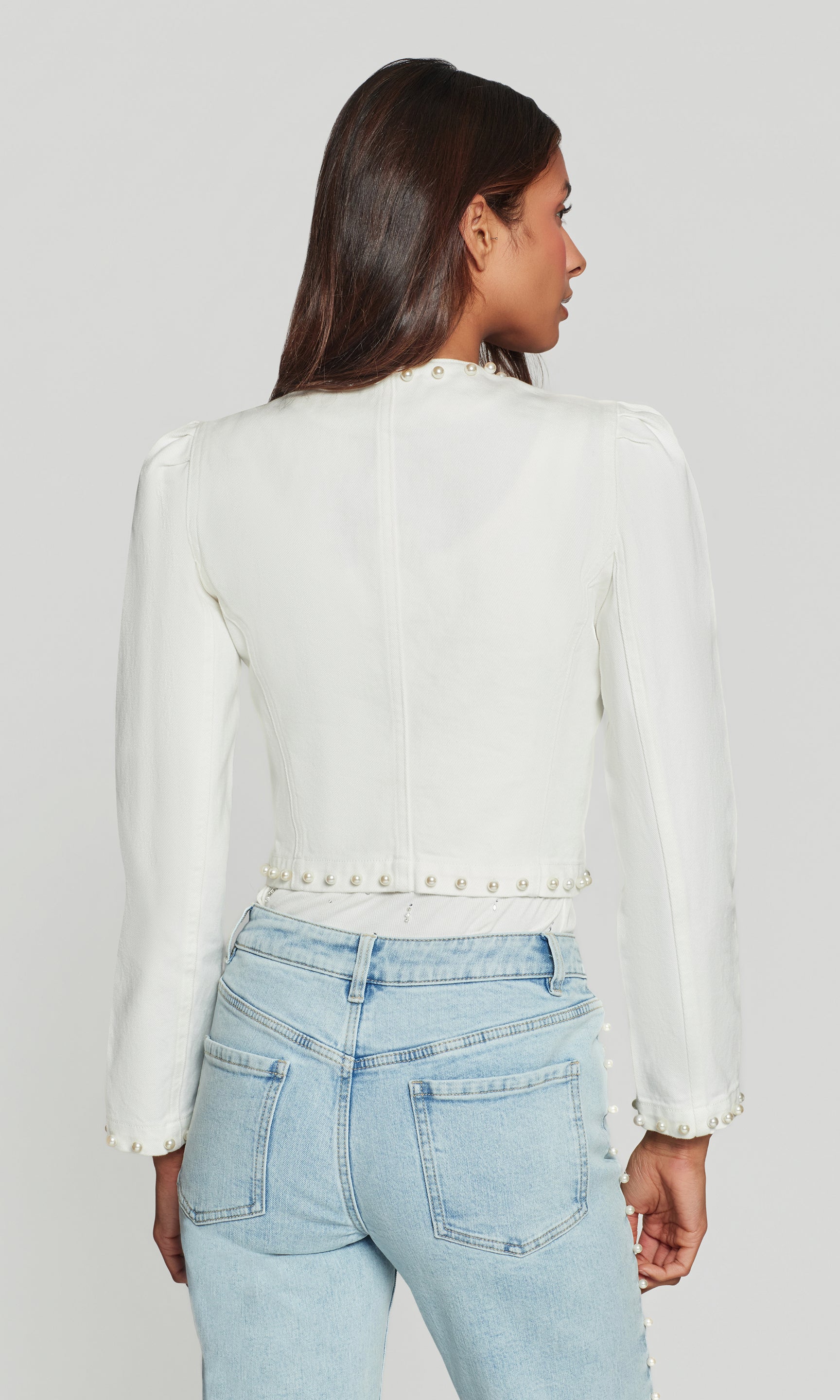 Person wearing a white jacket with pearl details and light blue jeans on a plain background
