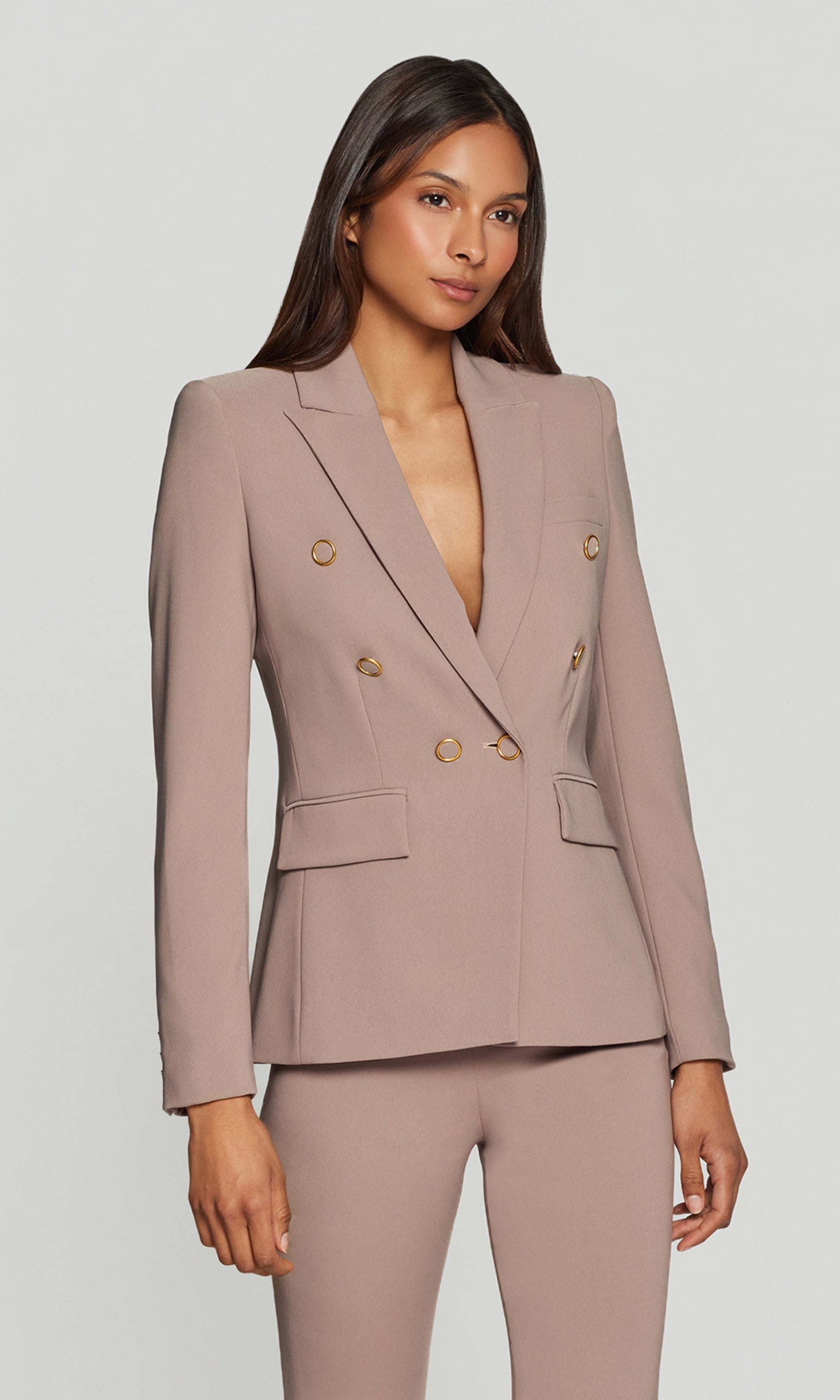 Side view of woman wearing a beige suit with gold buttons on a plain background