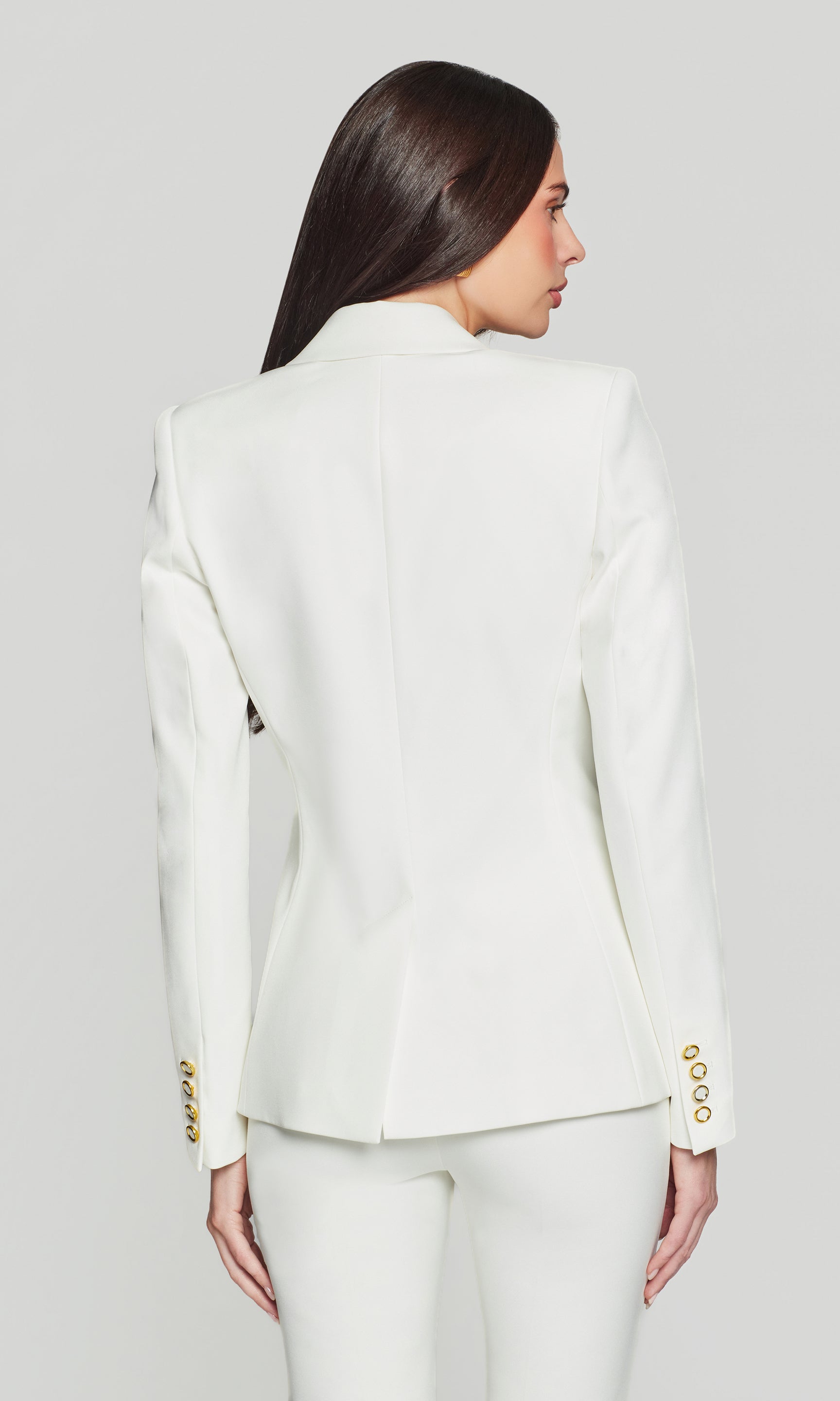 Back view of woman wearing a white suit with gold buttons on a plain background