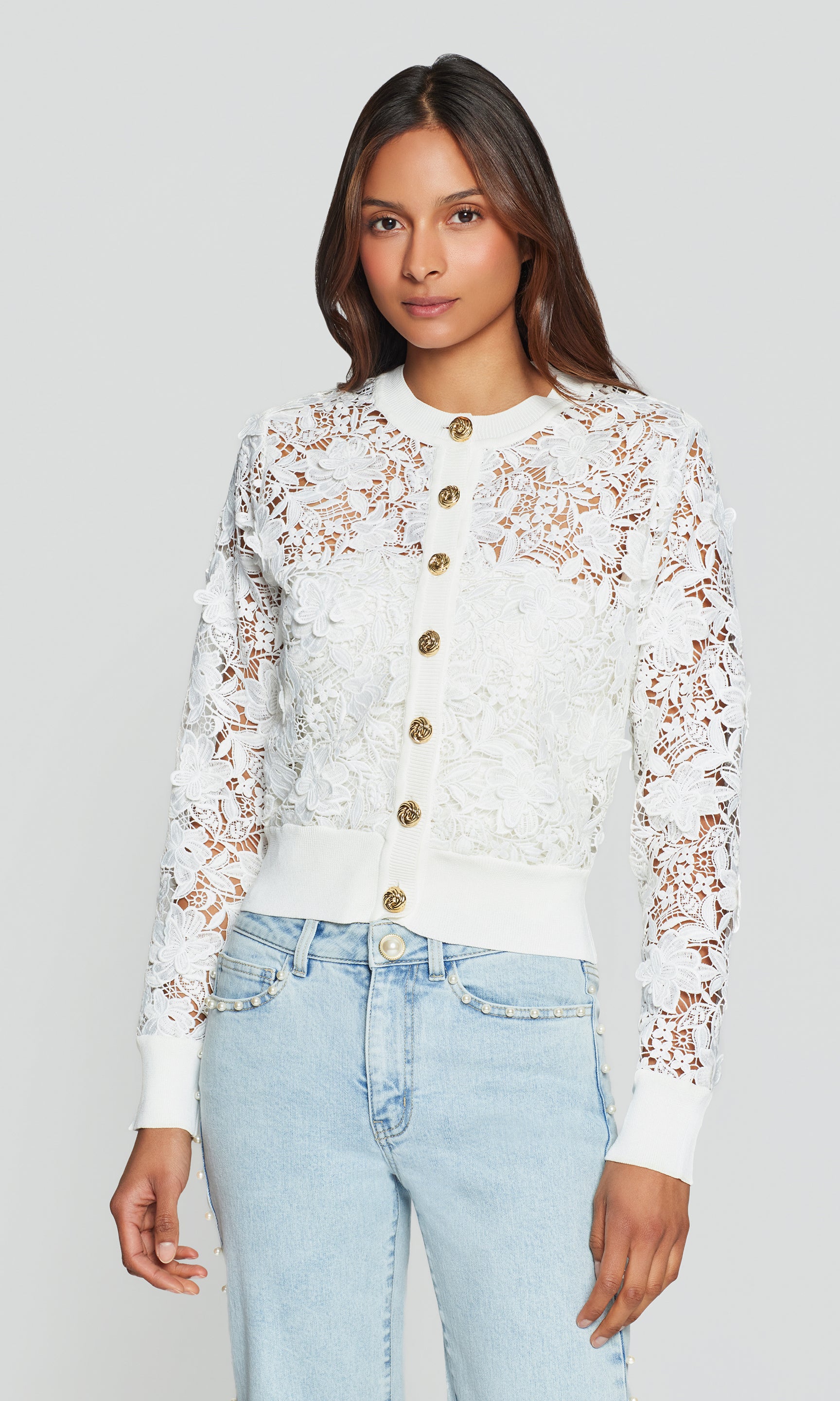 Woman wearing a white lace cardigan with gold buttons on a plain background