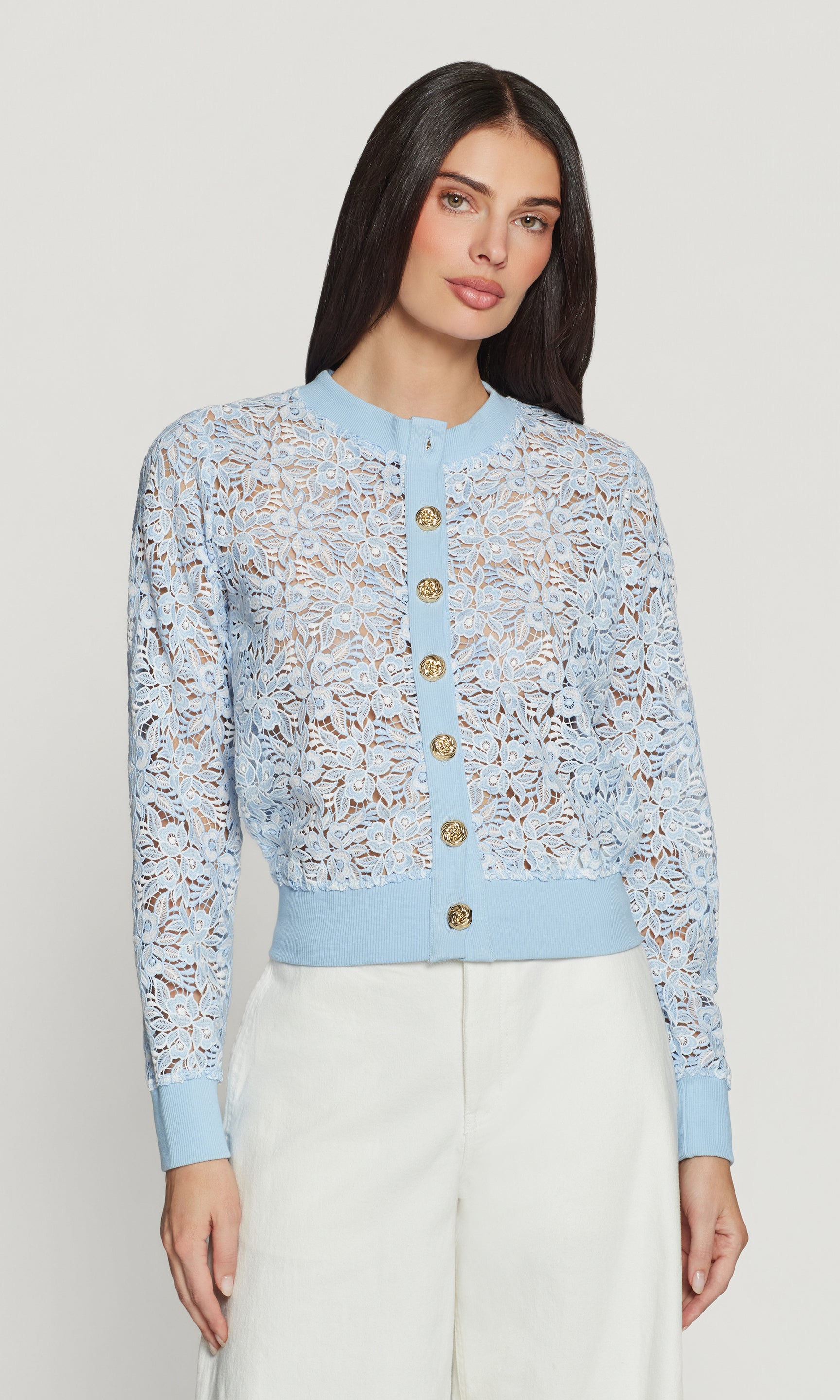 Woman wearing a light blue lace cardigan with gold buttons on a plain background