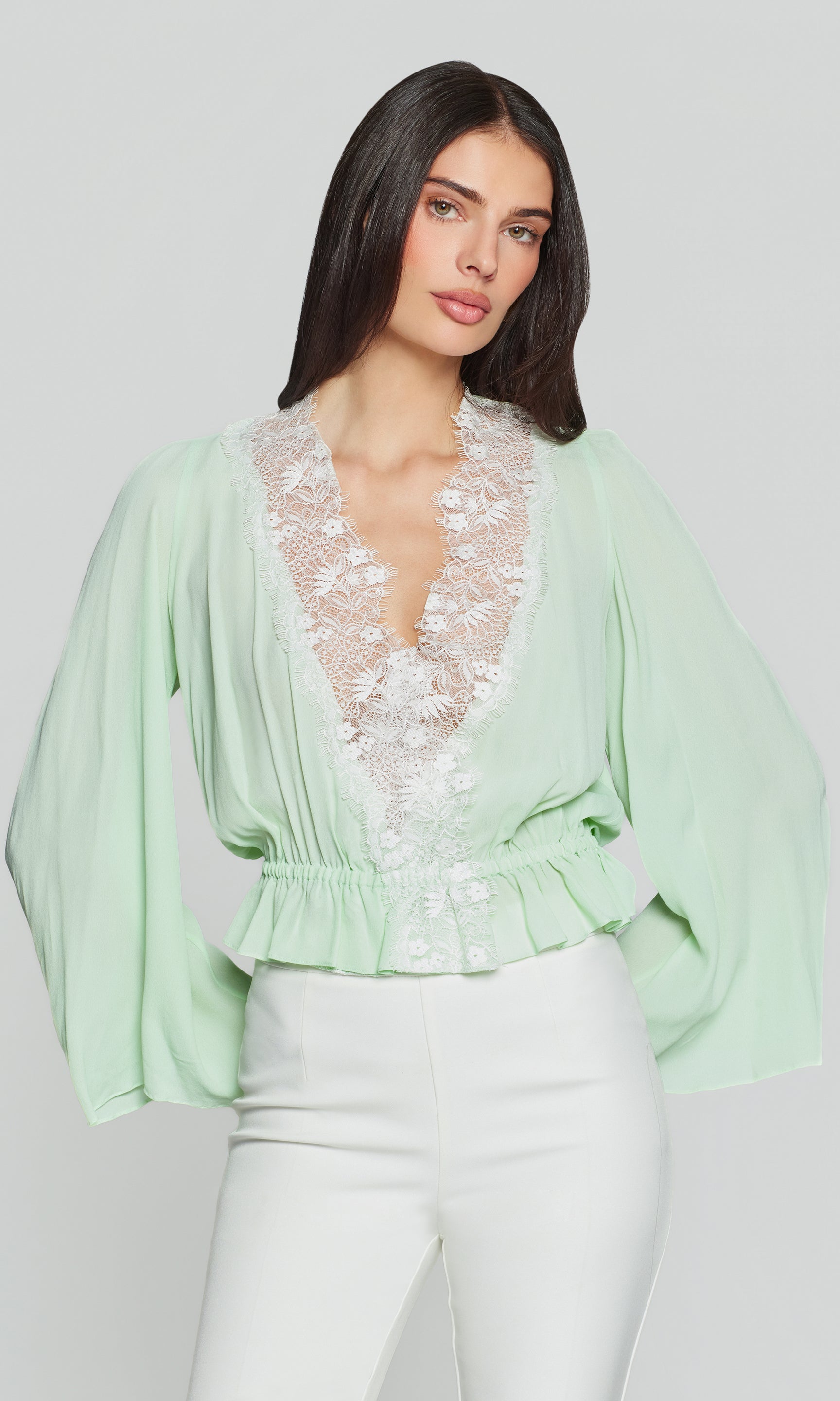 Woman wearing a light green blouse with lace detailing and white pants on a plain background
