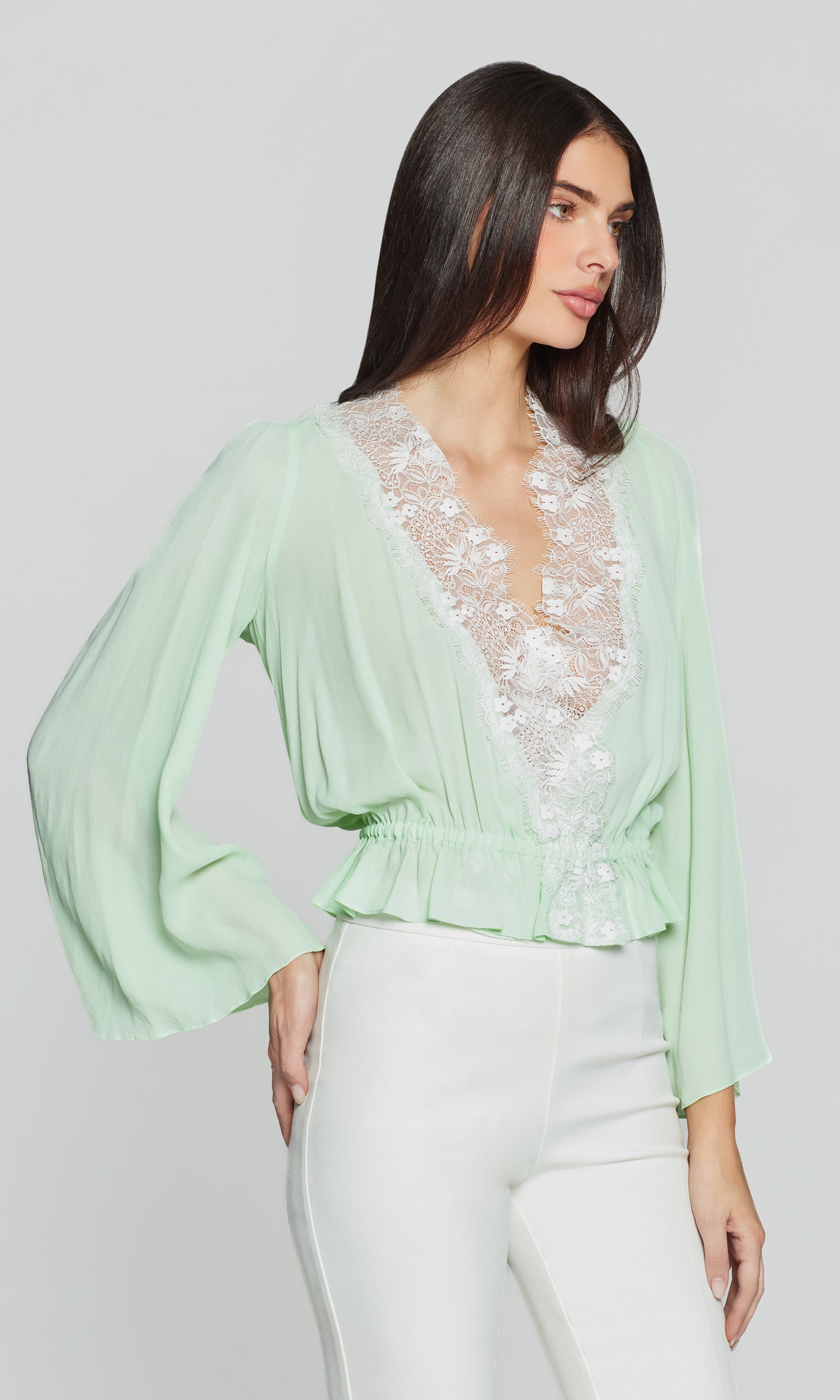 Side view of woman wearing a light green blouse with lace detailing and white pants on a plain background