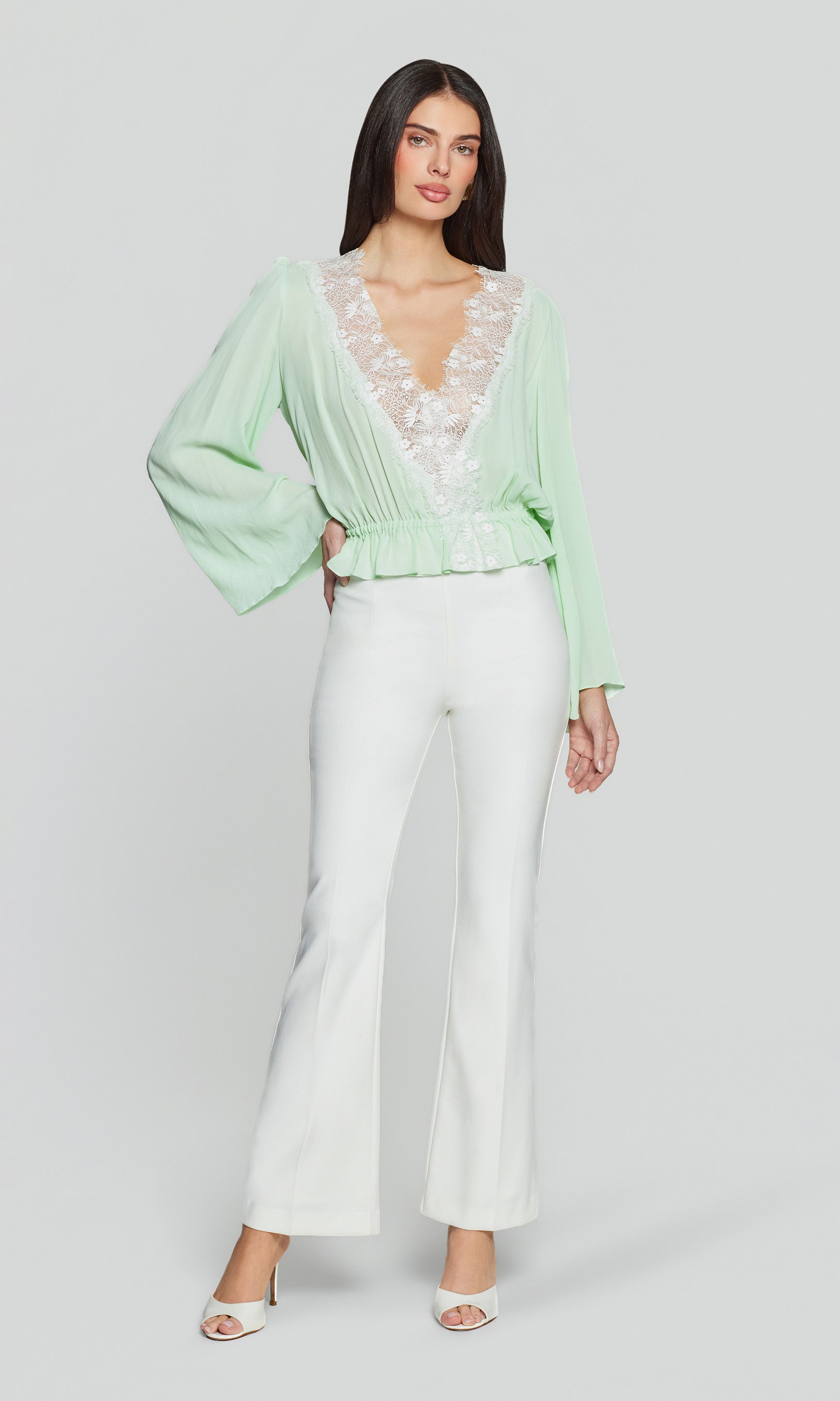 Woman wearing a light green blouse with lace details and white pants on a plain background
