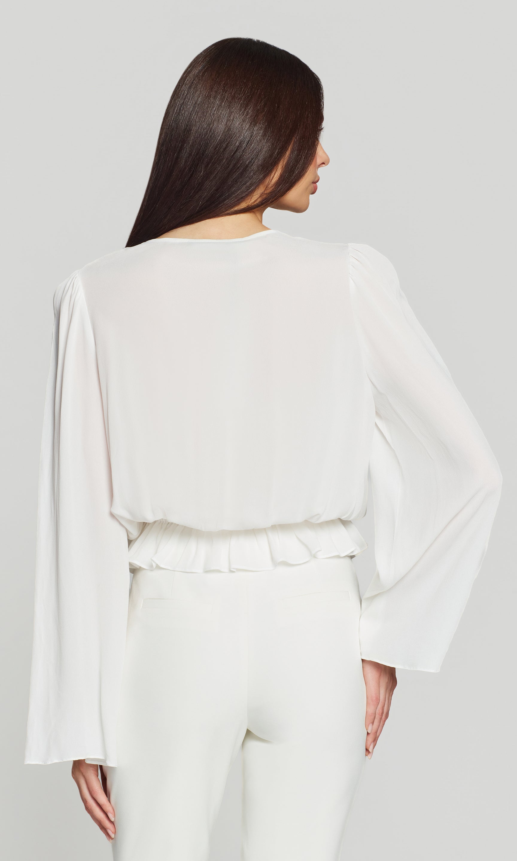 Back view of woman wearing a white flowy blouse and white pants against a plain background