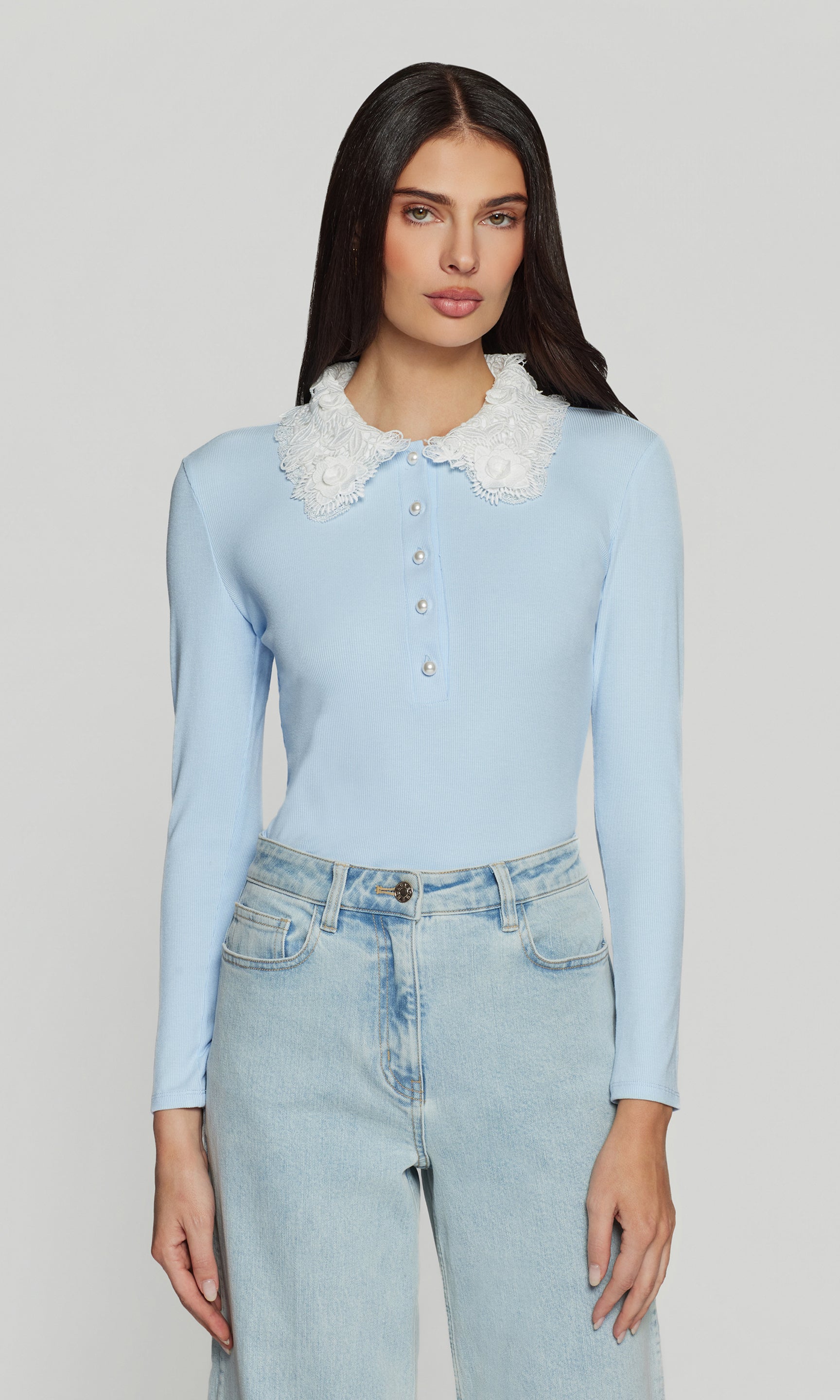 Woman wearing a light blue sweater with a white lace collar and light blue jeans on a plain background