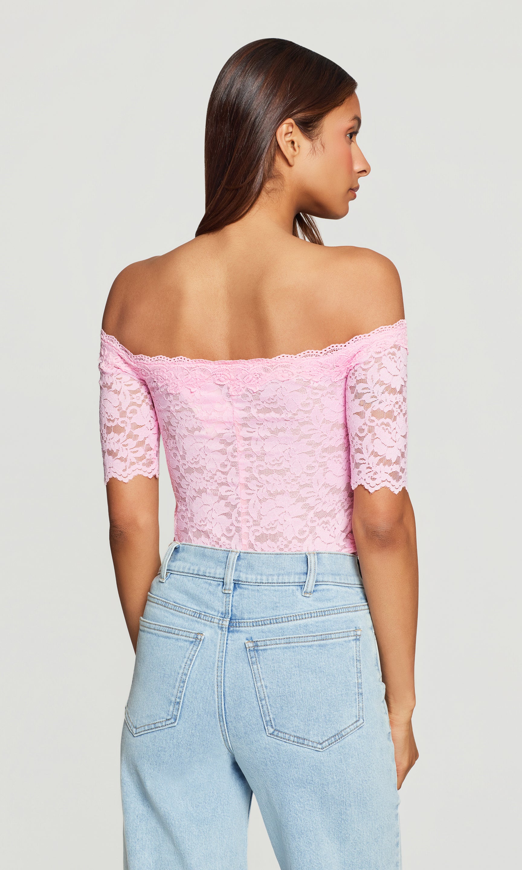Back view of woman wearing a pink lace off-shoulder top and light blue jeans on a plain background
