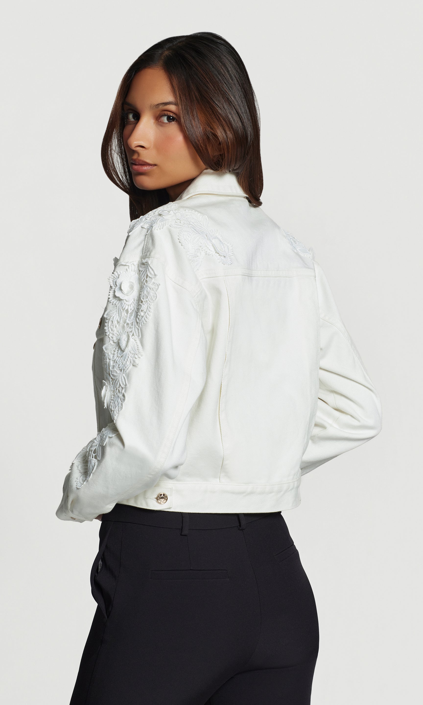 Back view of woman wearing a white jacket with lace details on a plain background