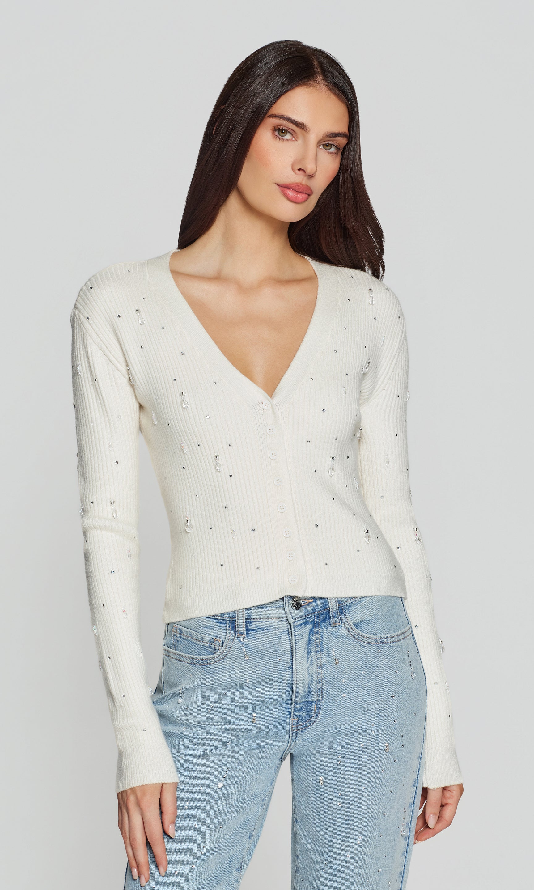 Woman wearing a white long-sleeve top with crystals and light blue jeans on a plain background