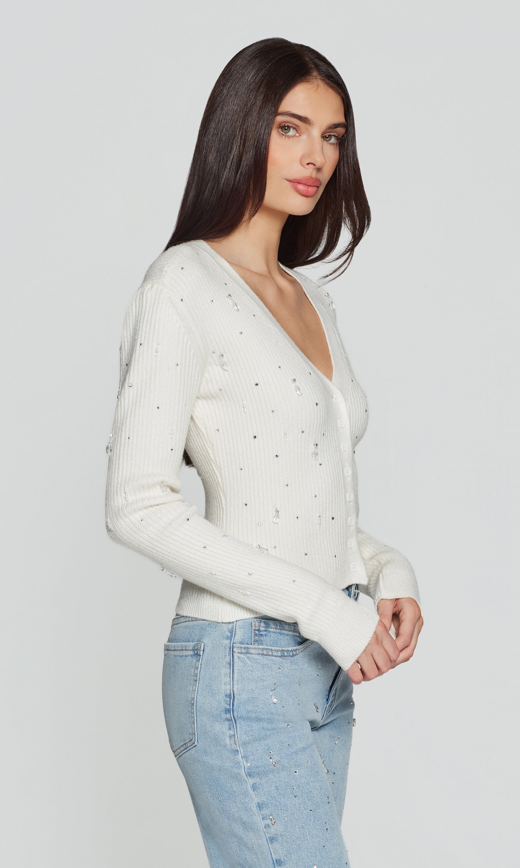 Side view of woman wearing a white cardigan with crystal embellishments on a plain background