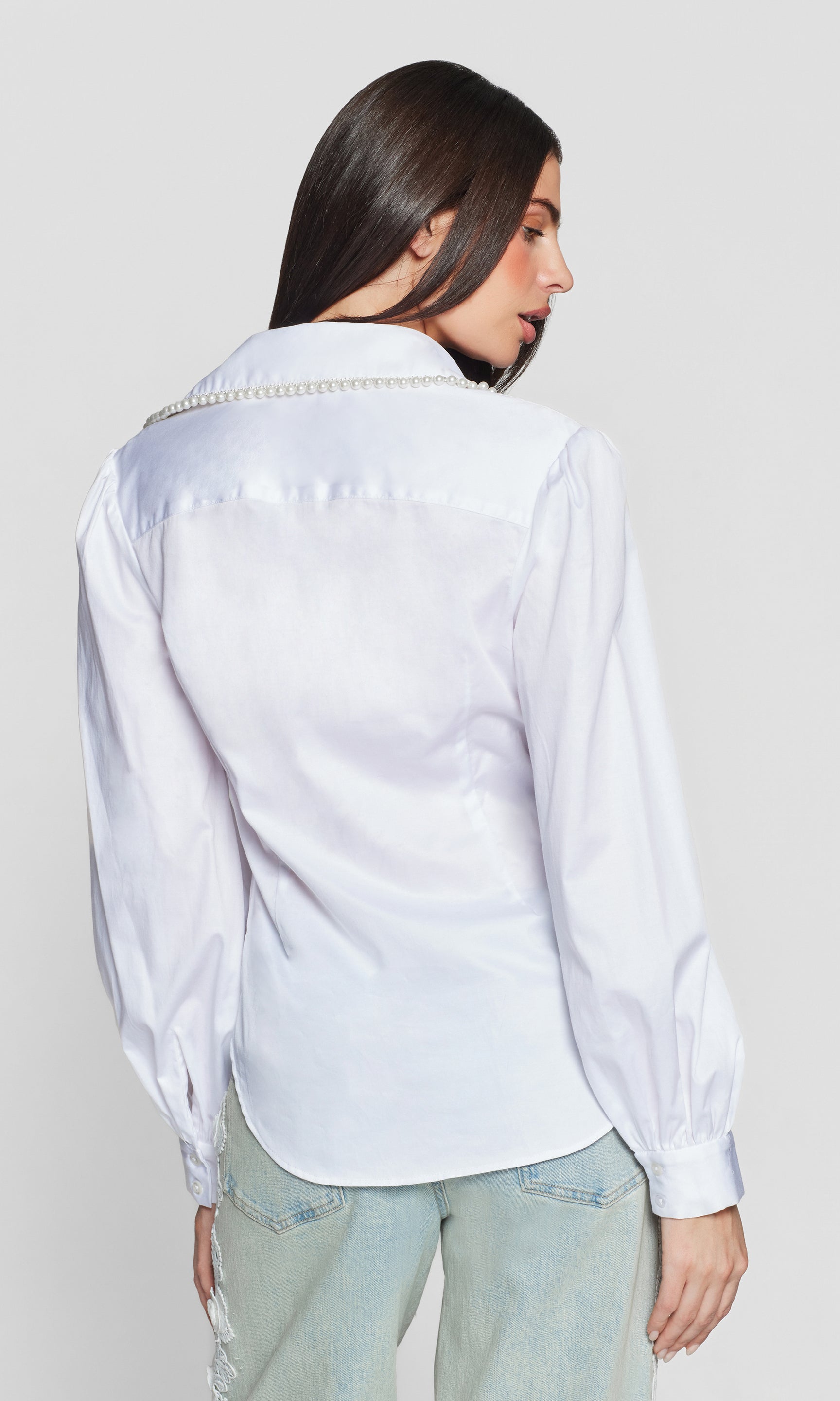 Back view of woman wearing a white blouse with decorative pearl trim on a plain background