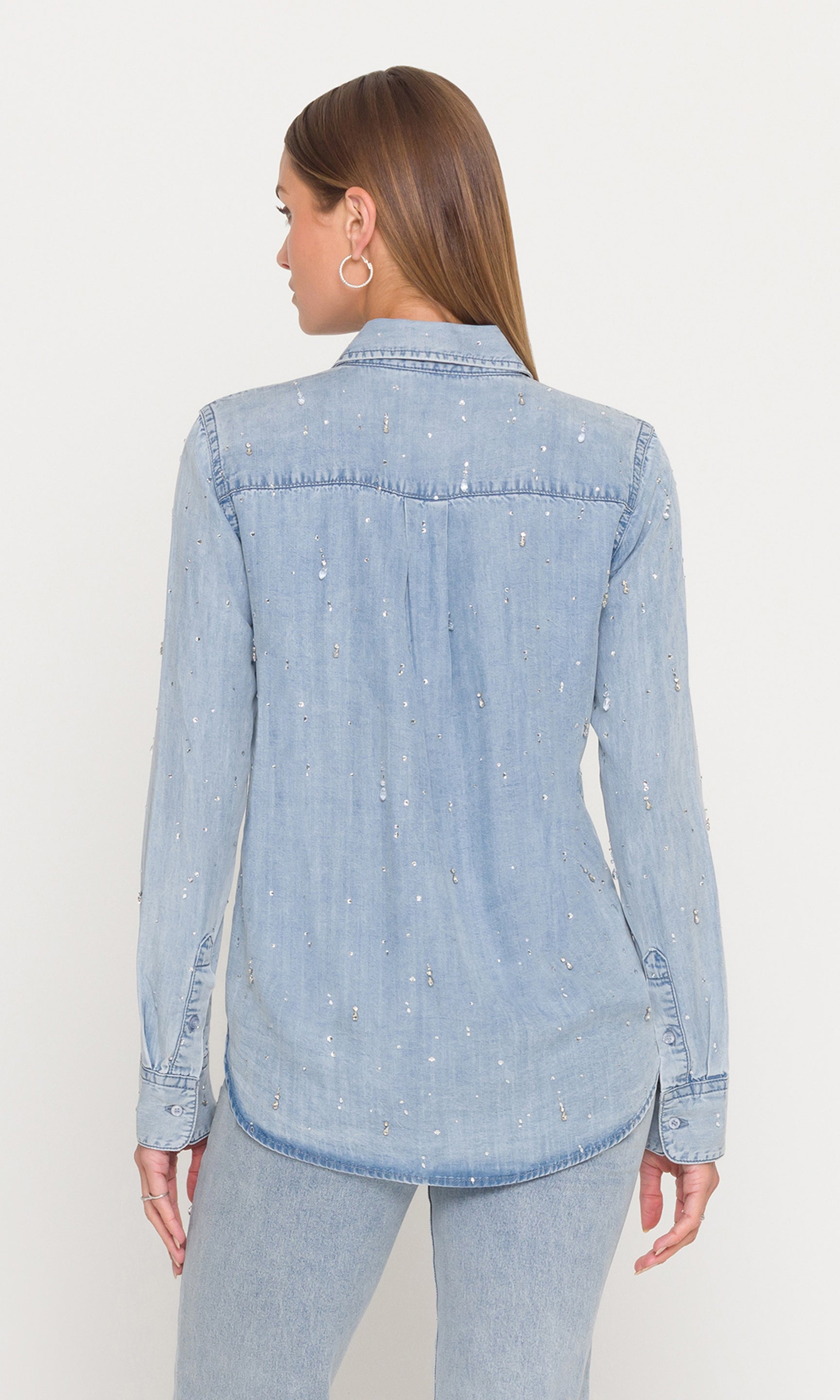 Back view of woman wearing a light blue denim shirt with crystals on a white background