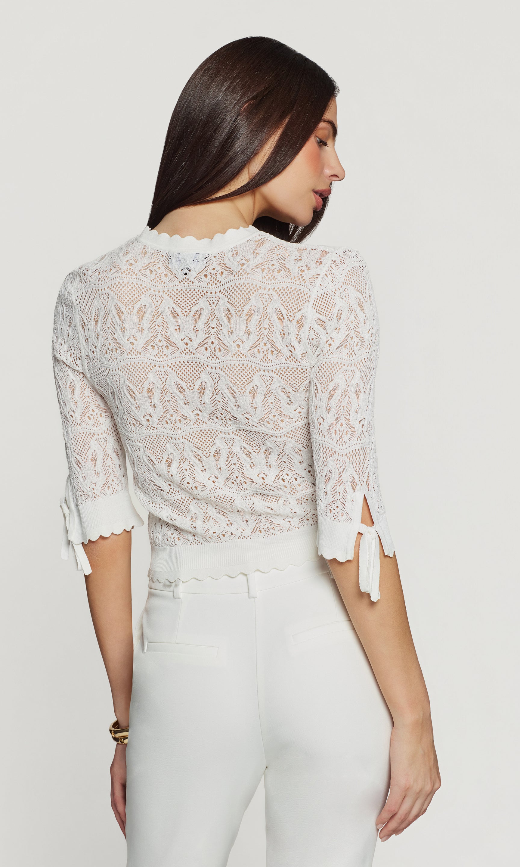 Back view of woman wearing a white lace cardigan with butterfly knit detail on a plain background
