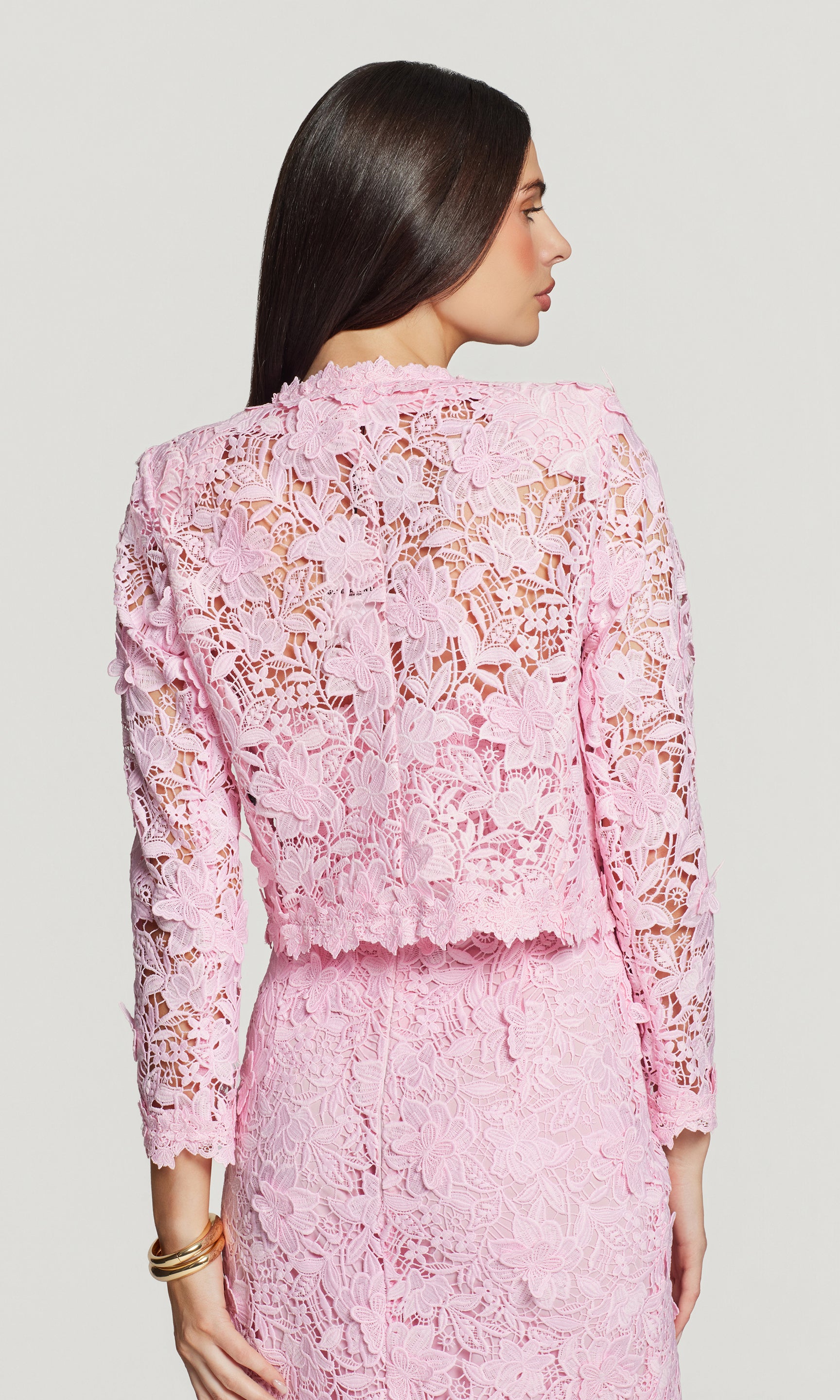 Back view of woman wearing a pink lace jacket over a matching dress against a light gray background