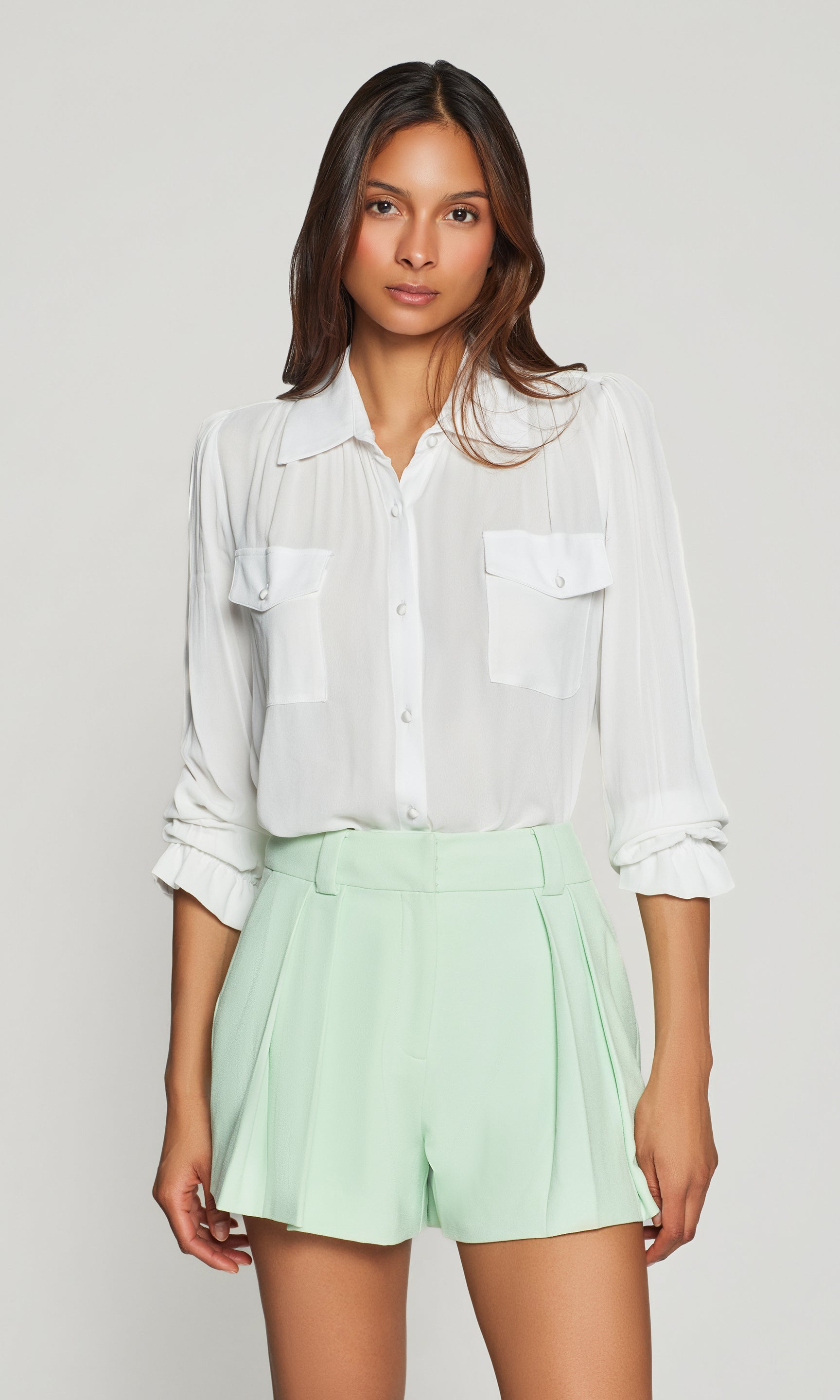 Model wears the Priya Georgette Shirt in white in a front view, showing the airy drape, button-front detail, and flap chest pockets styled with mint shorts