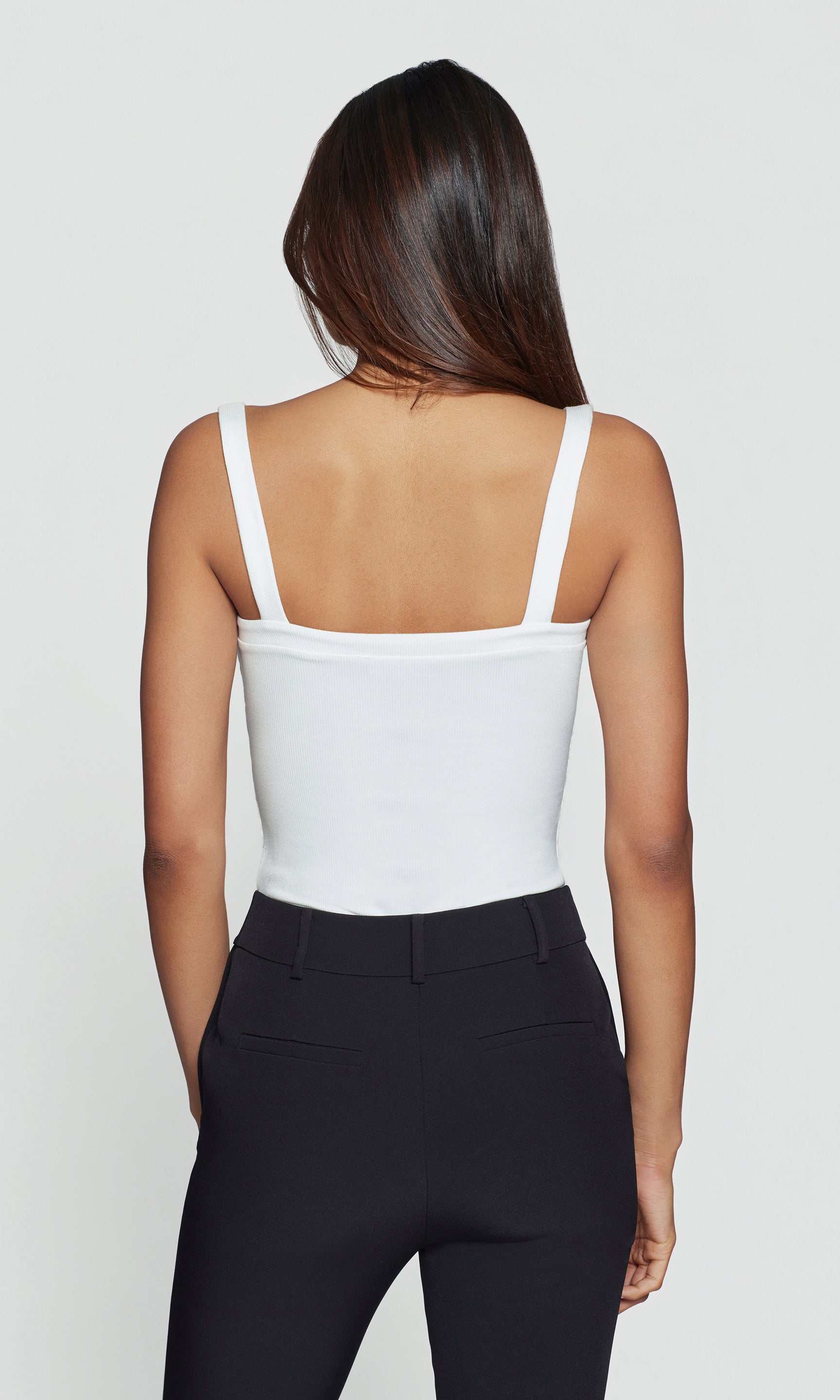 Back view of person wearing a white tank top and black pants on a plain background