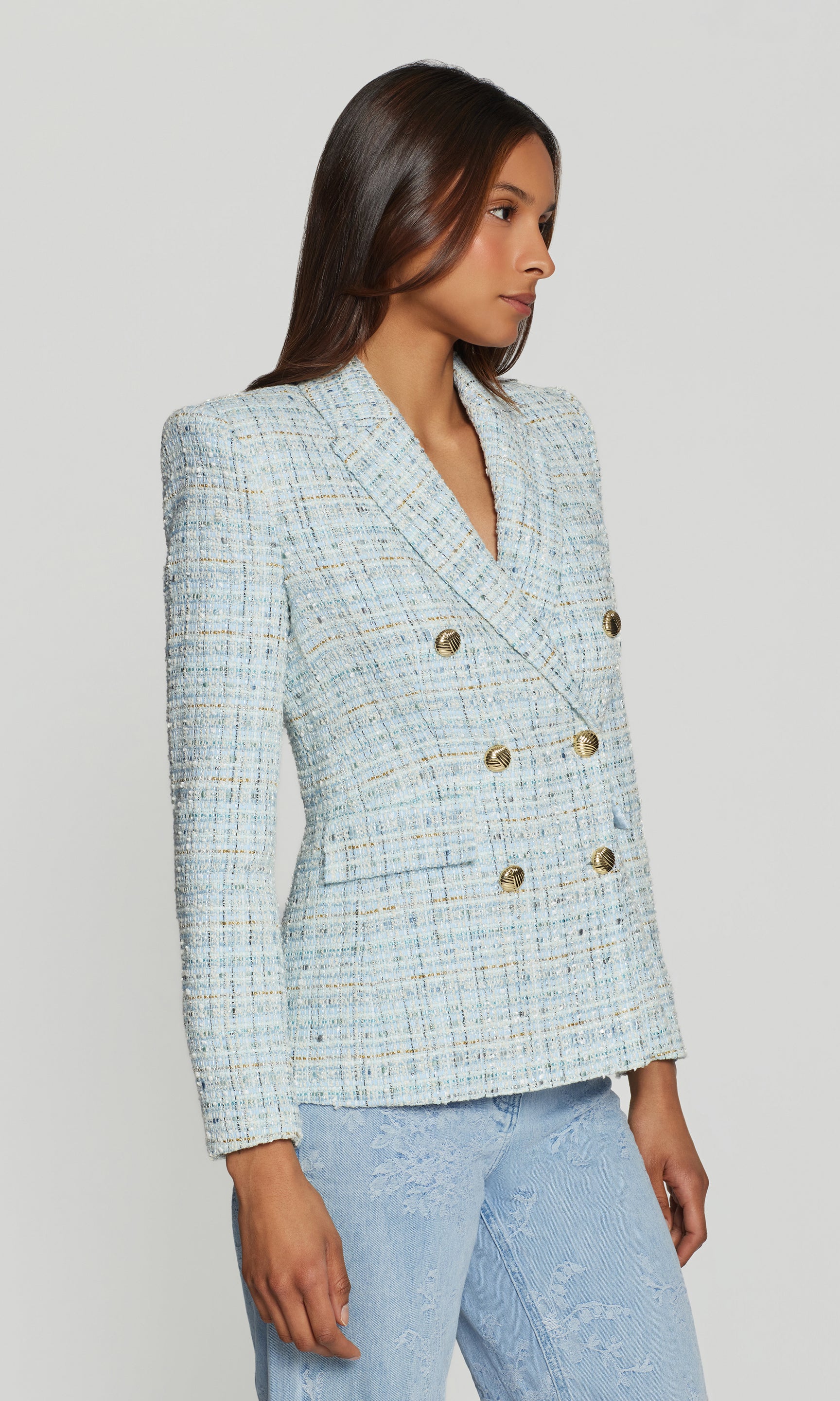 Side view of woman wearing a light blue tweed blazer with gold buttons on a white background