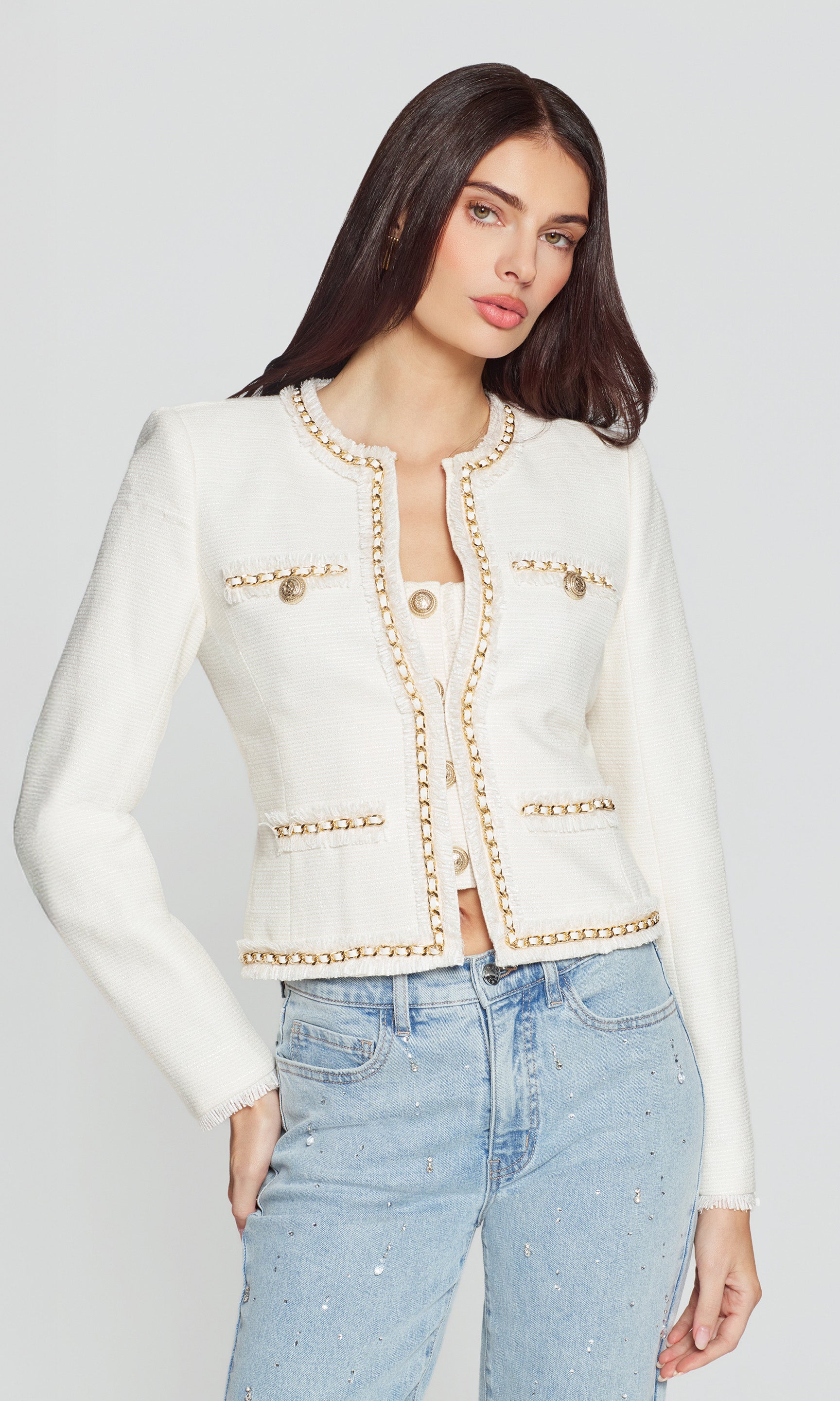 Woman wearing a white jacket with gold accents and light blue jeans on a plain background