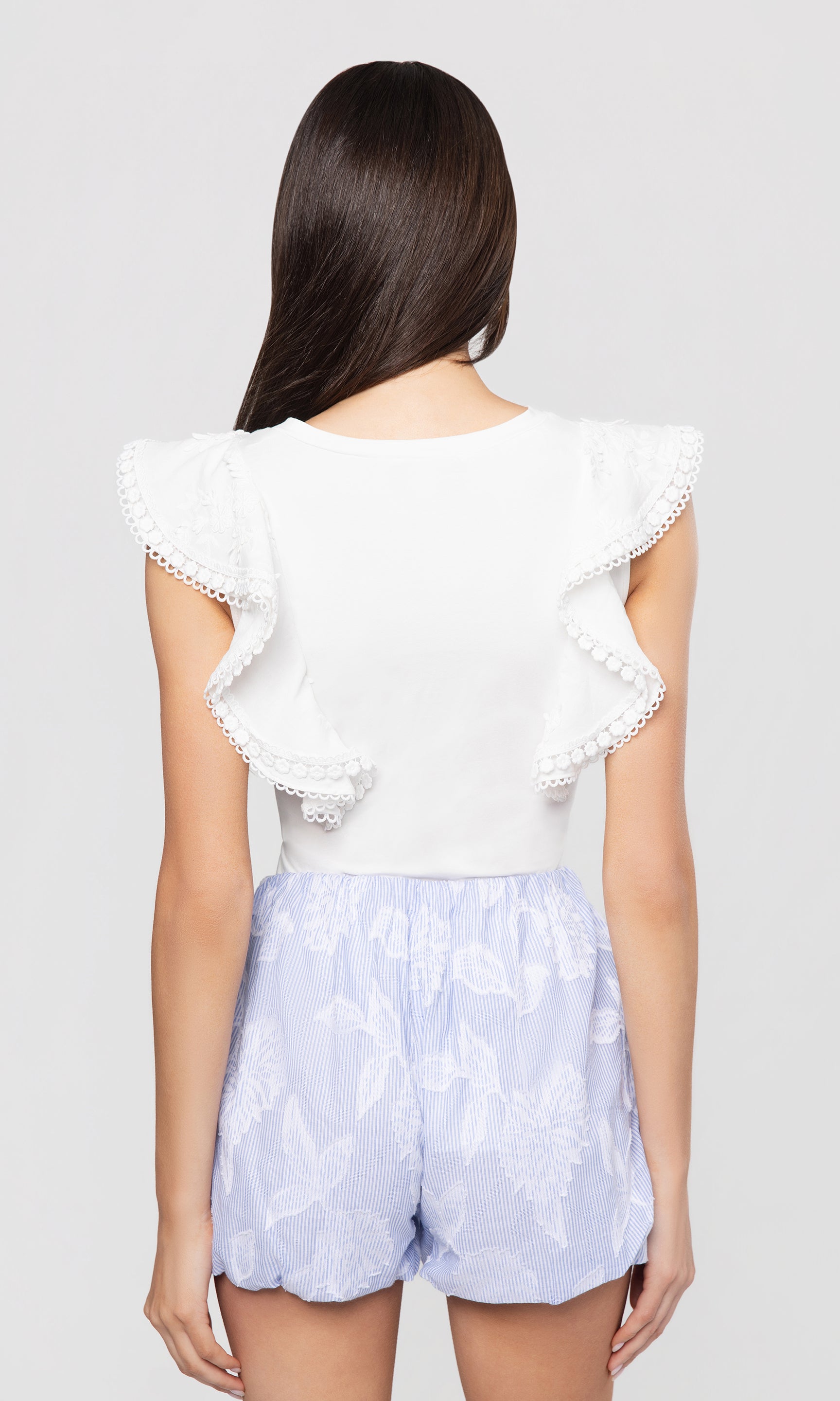 Back view of woman wearing a white top with ruffled sleeves and baby blue shorts on a white background