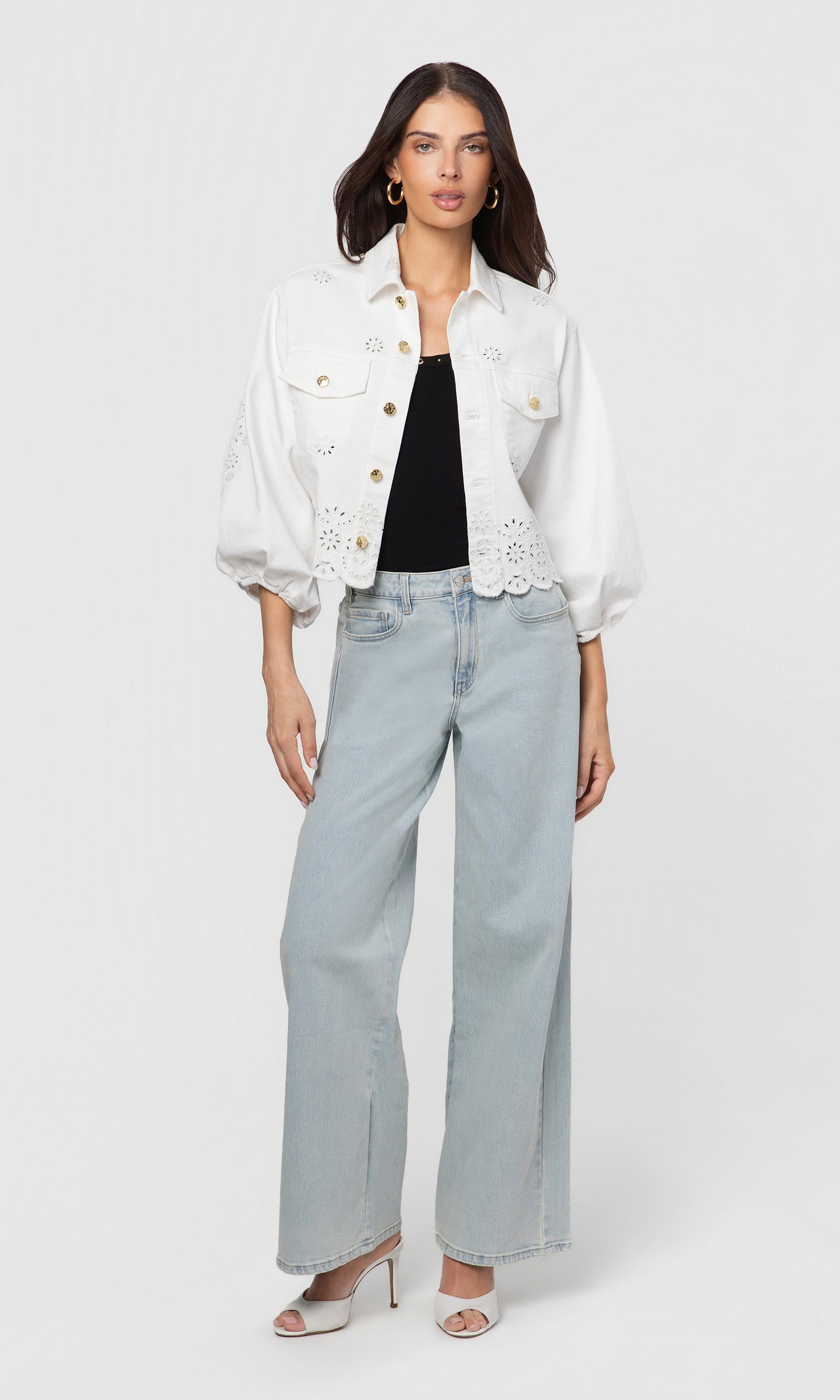 Woman wearing a white jacket with lace details, black top, and light blue jeans on a white background