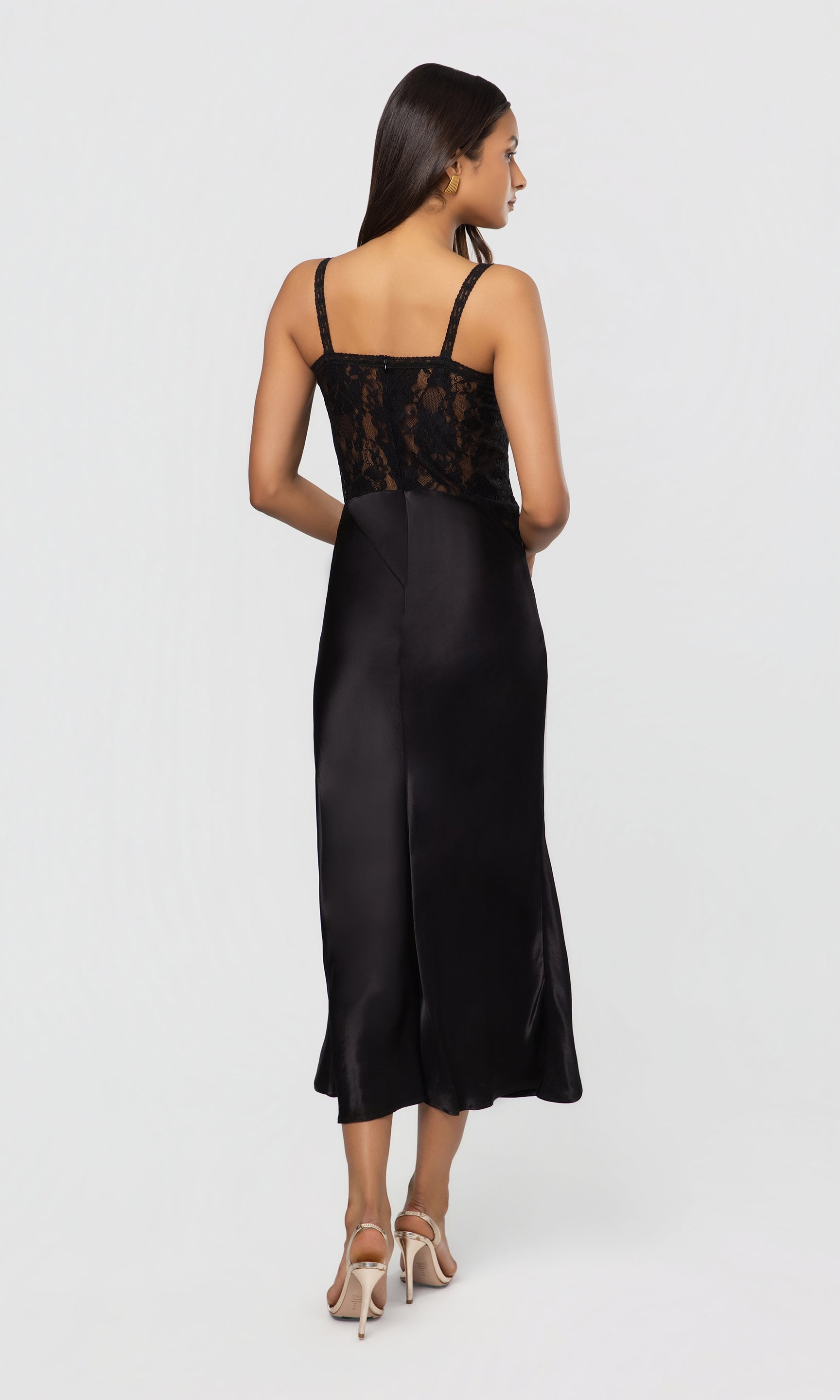 Back view of woman wearing a black satin slip dress with lace detailing on a white background