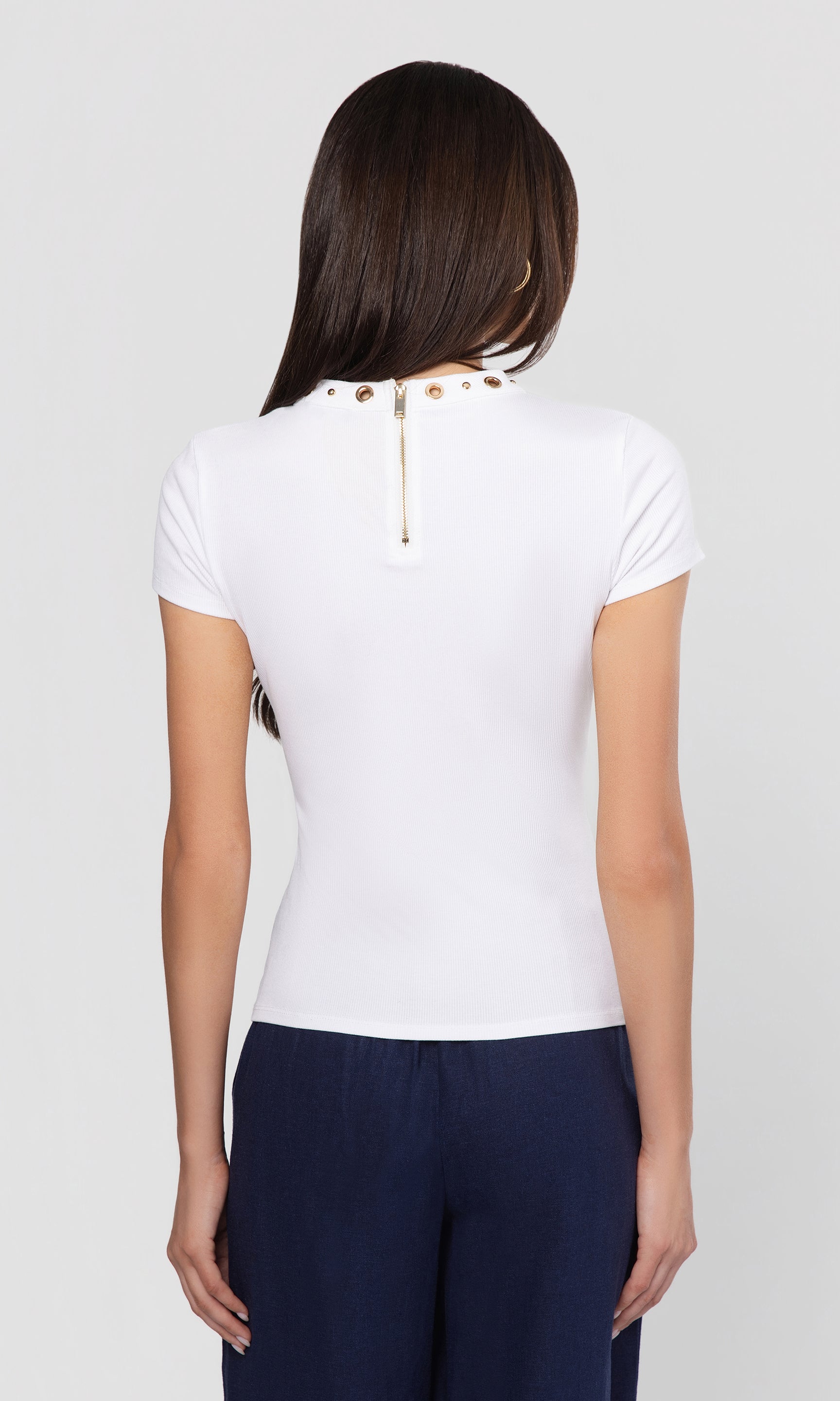 Back view of woman wearing a white top with grommet details on the neckline on a plain background.