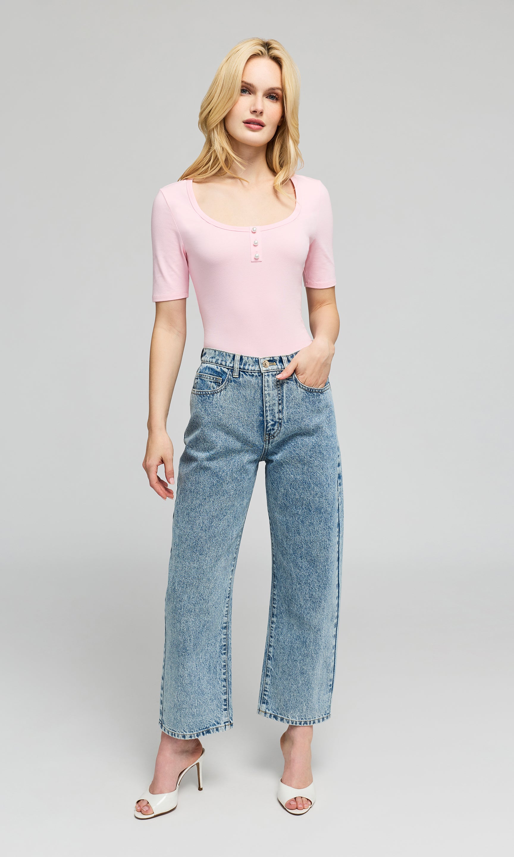 Model wearing a light pink top and blue jeans, paired with white heeled sandals, on a plain background.