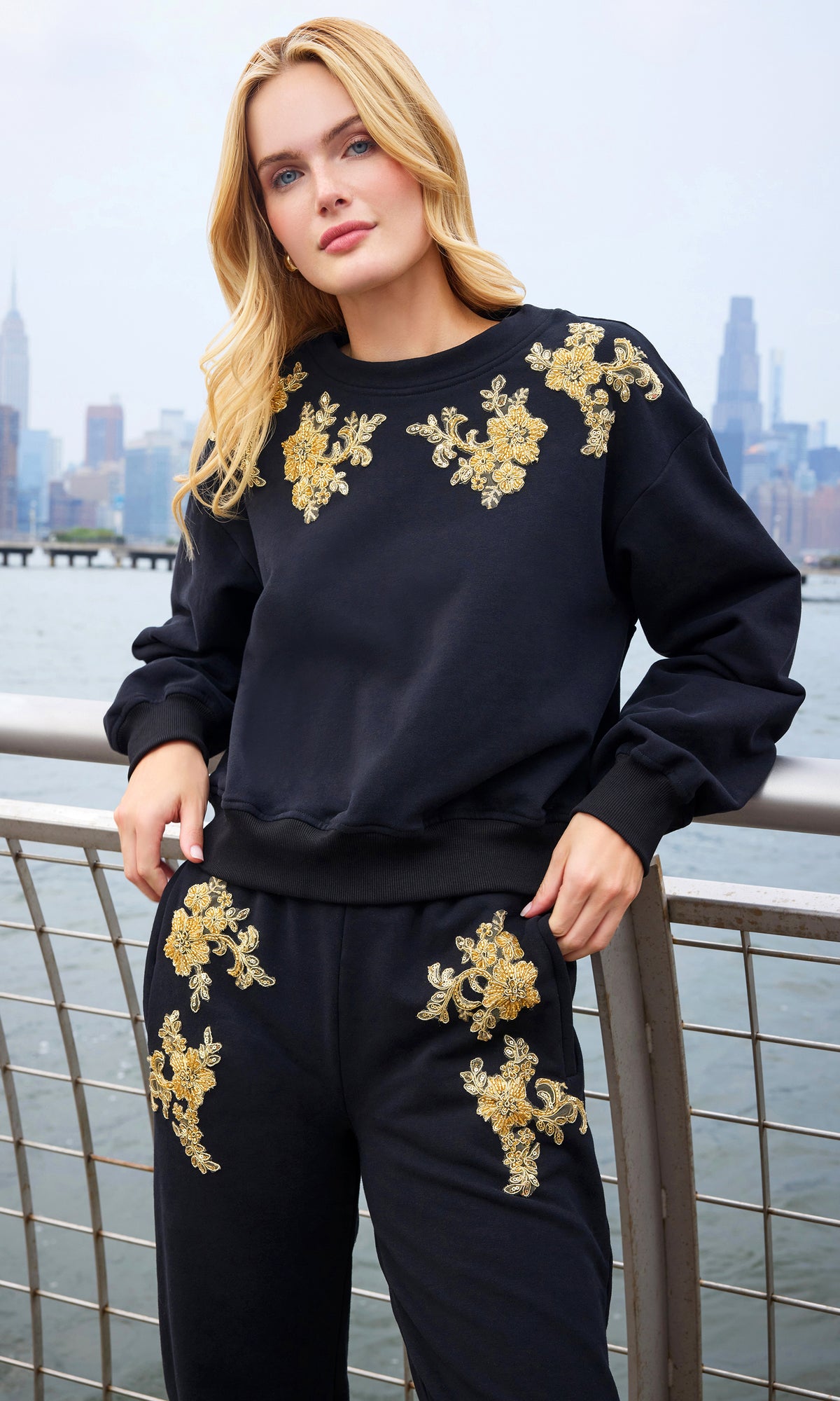 A woman wearing a matching black sweatshirt and jogger pants set with ornate gold floral embroidery, posed outdoors with a city skyline and water in the background.