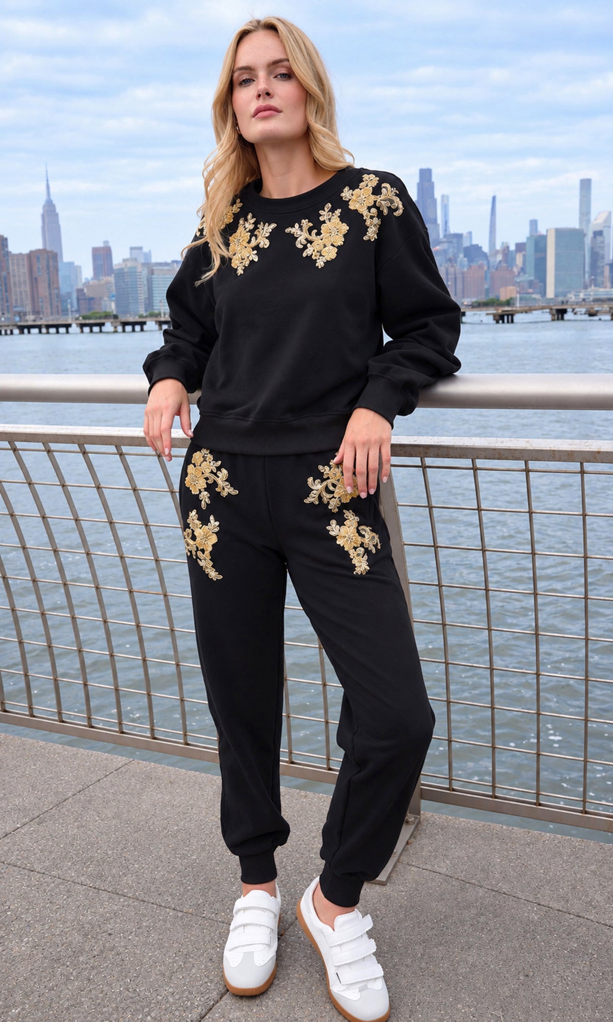 A woman stands outdoors by a waterfront railing wearing a black sweatshirt and jogger pants set with gold floral embroidery, paired with white sneakers.