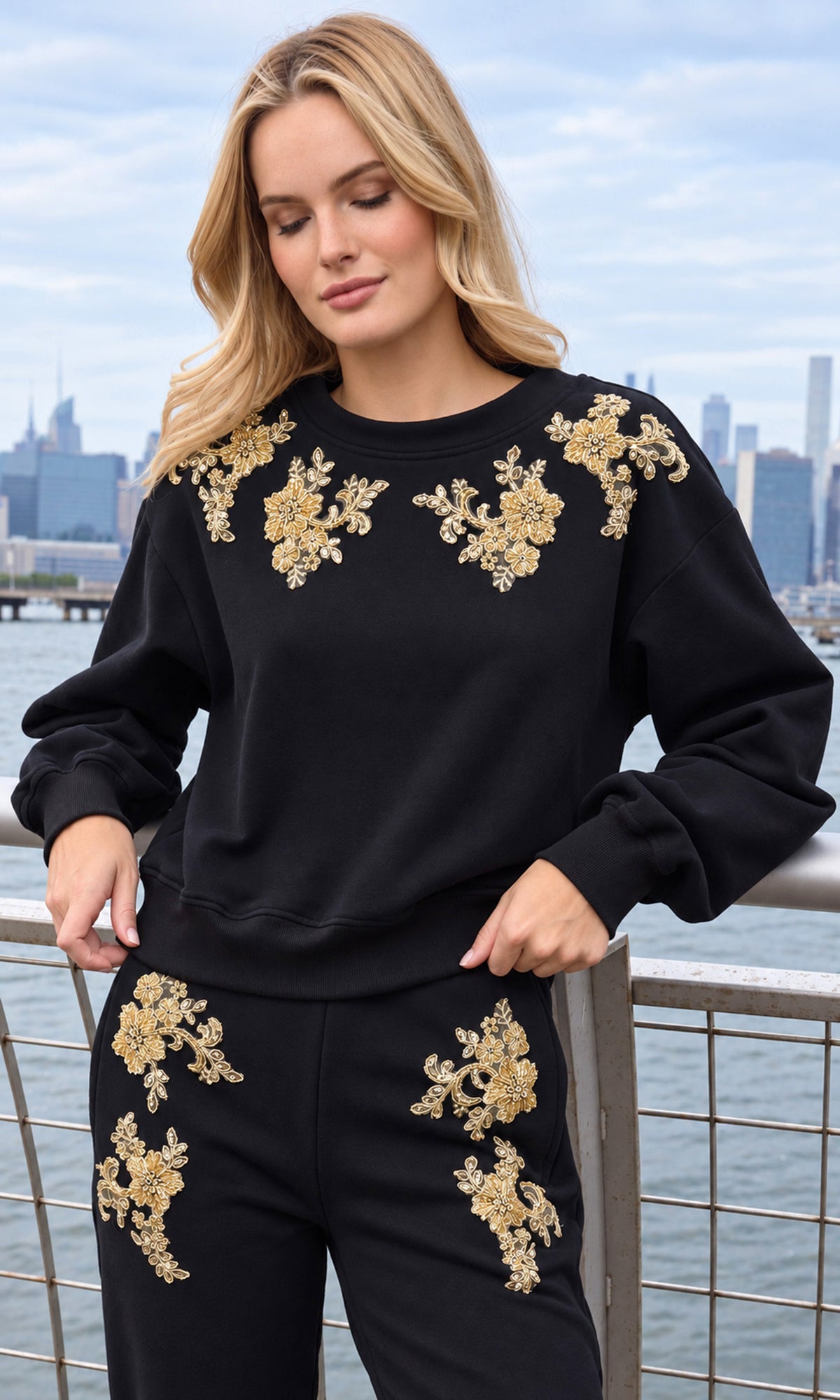 A woman wearing a black crewneck sweatshirt and matching sweatpants with gold floral embroidery, standing outdoors with a city skyline and water in the background.