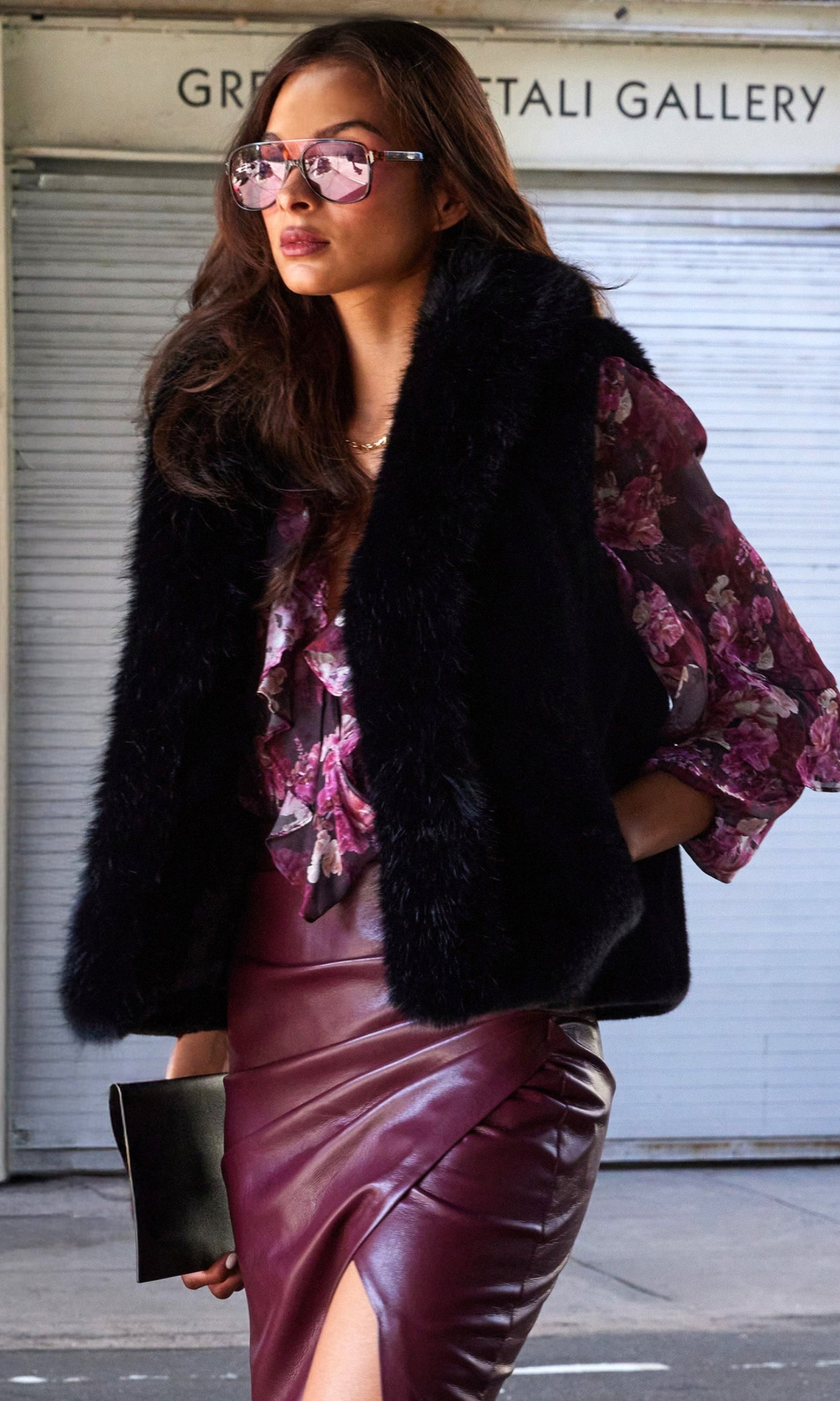 Woman wearing a black fur vest, floral blouse, and burgundy leather skirt in an urban setting.