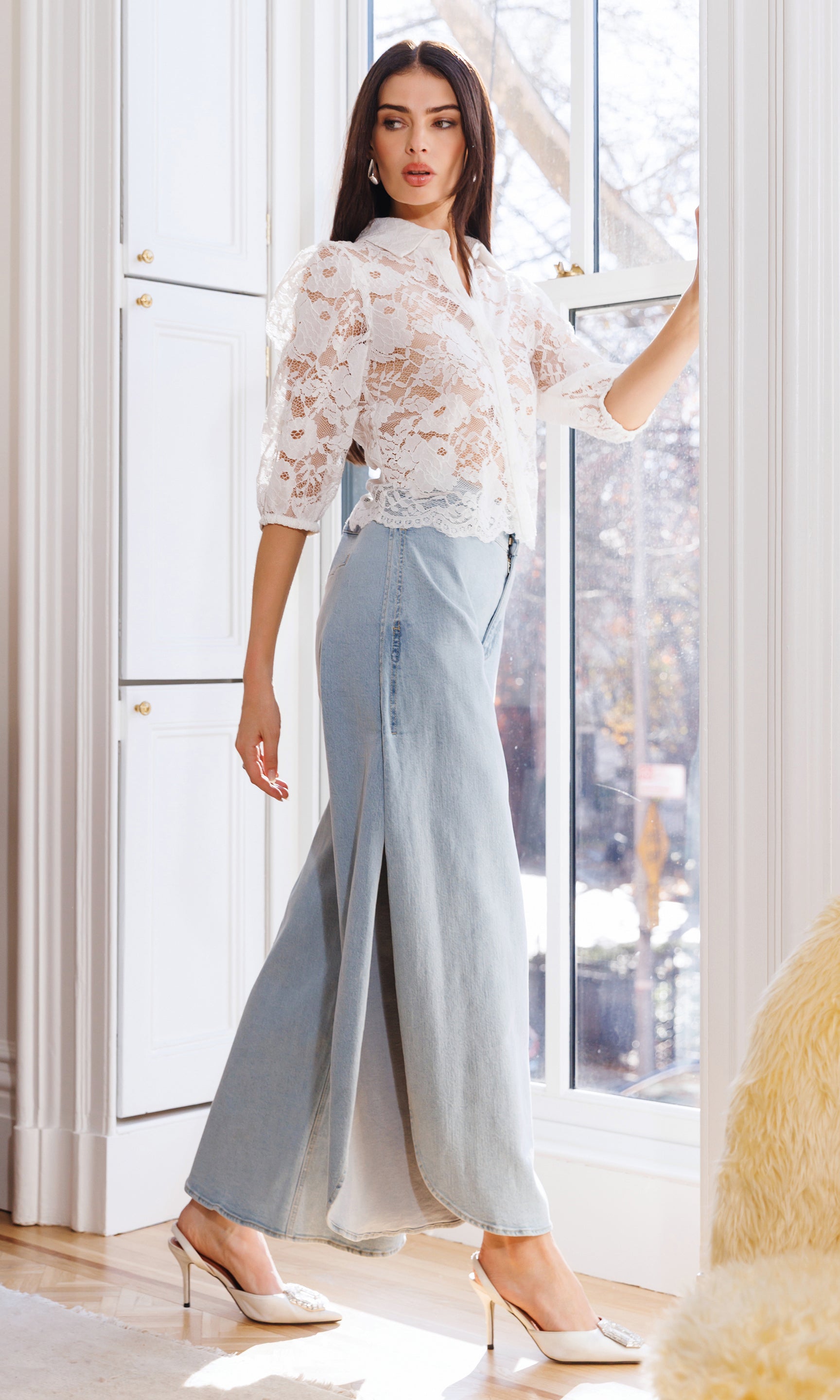 Woman wearing a white lace top and light blue pants in a bright room.