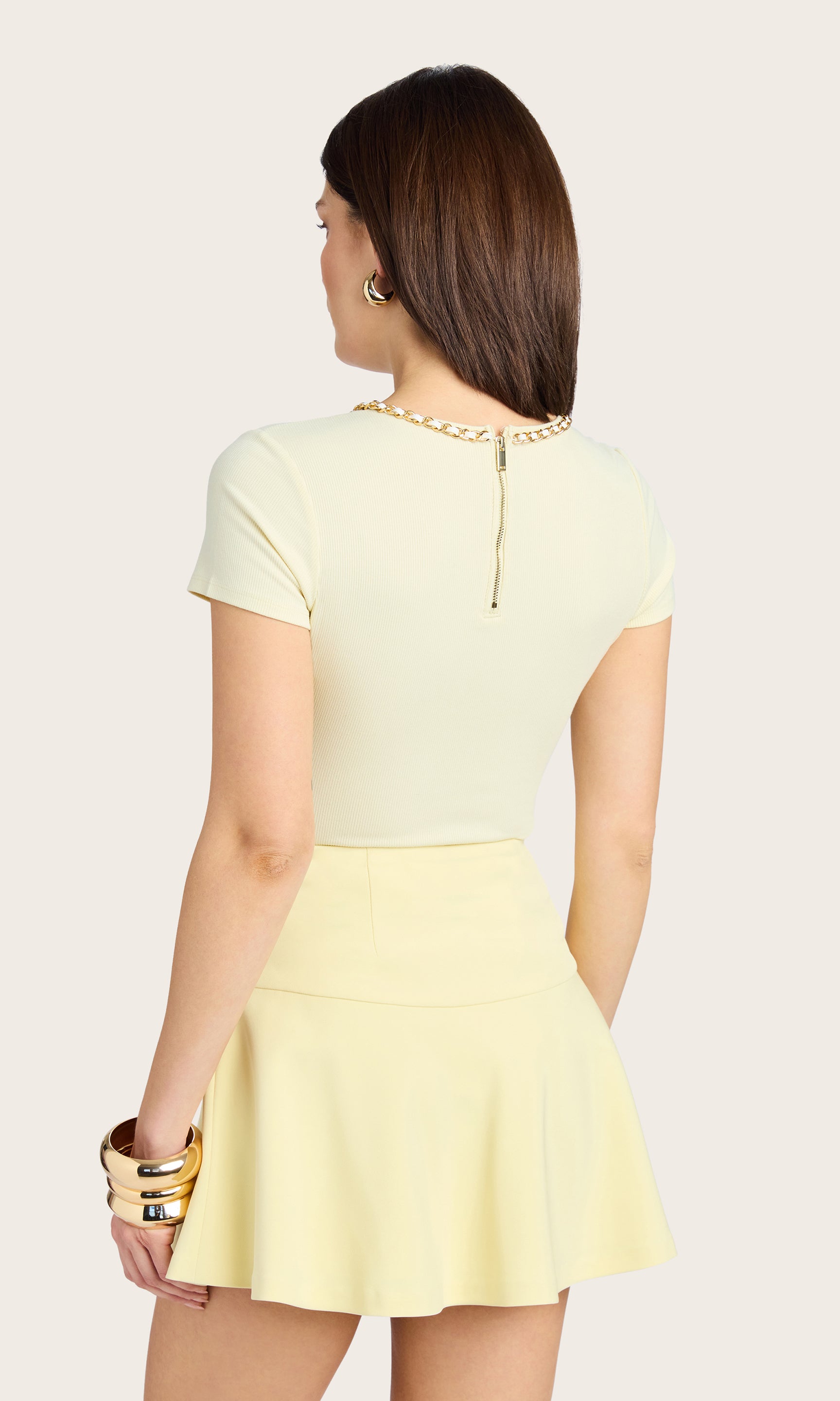 back view of model wearing the Tate Chain Top in pastel yellow, paired with a pastel yellow skirt, and accessorized with a gold bracelet and earrings