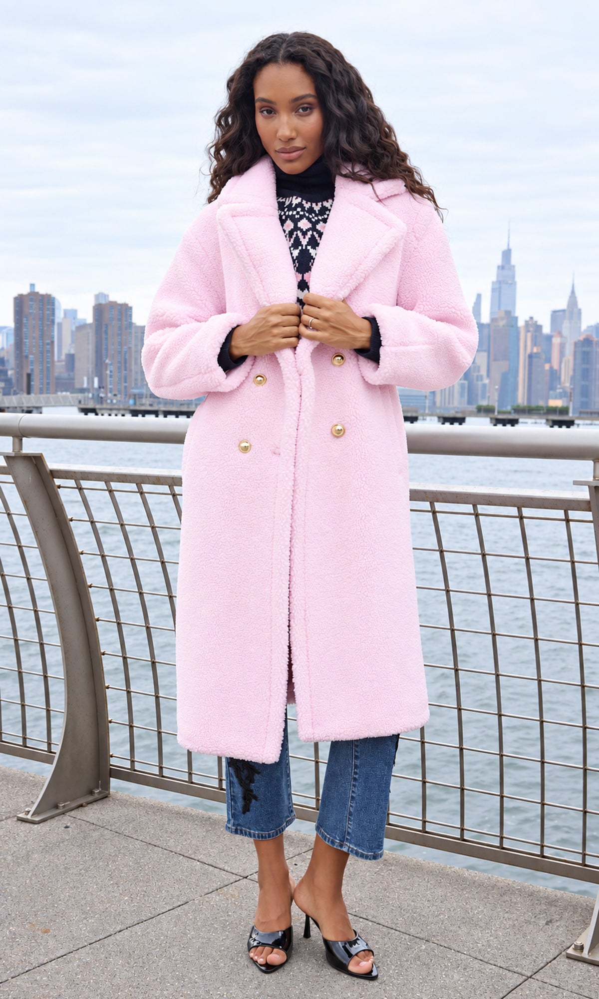 A woman wearing a light pink fuzzy double-breasted coat with gold buttons, styled over a black patterned top and blue jeans, standing on a waterfront with a city skyline in the background.