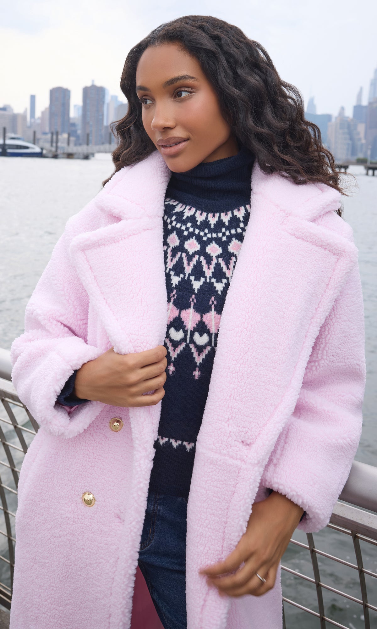 A woman wearing a light pink teddy coat over a navy and pink patterned sweater, standing outdoors near a waterfront railing with a city skyline in the background.