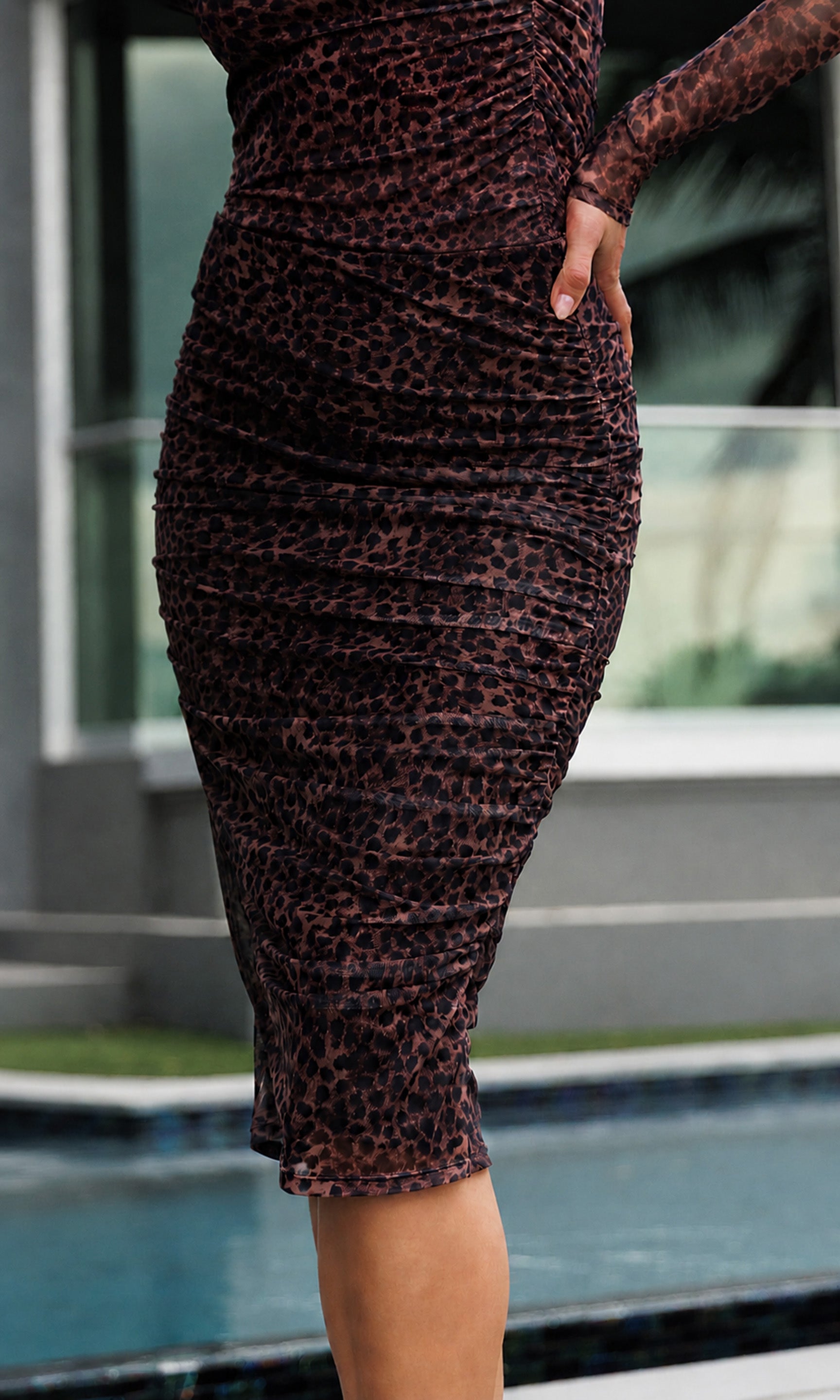 A woman wearing a fitted leopard-print midi skirt and matching top, standing outdoors near a pool and modern building background.
