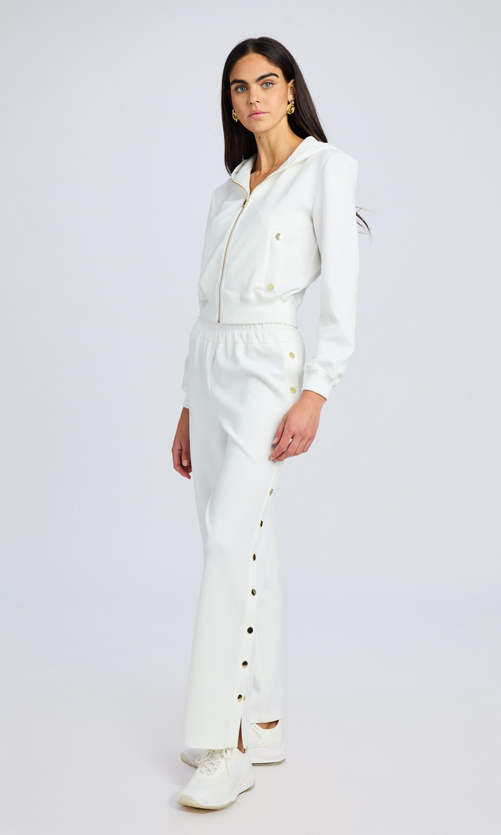 model stands wearing a white scuba fabric zip up hoodie with matching white pants featuring gold snaps down the sides.