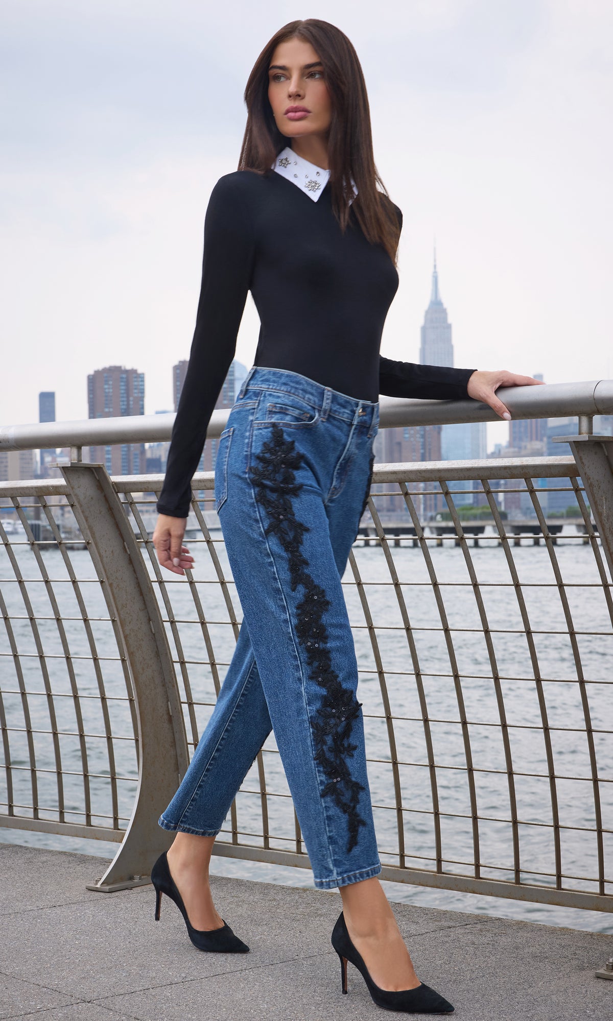 Model wearing a fitted black long-sleeve top with a white patterned collar, blue cropped jeans with black distressed embroidery down the side.