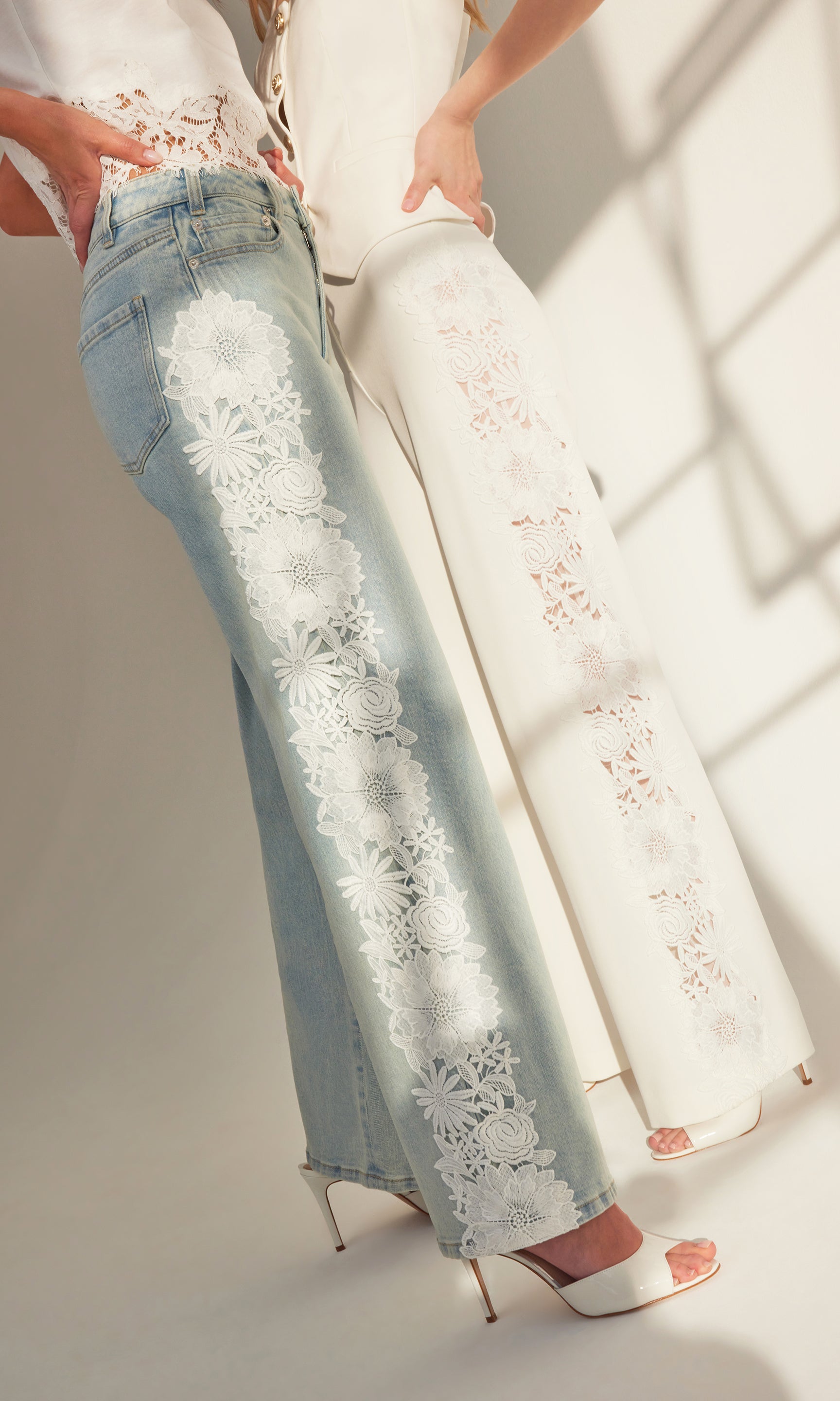 two models standing front to front both wearing pants that have lace details down the side of the pant legs, both paired with white tops