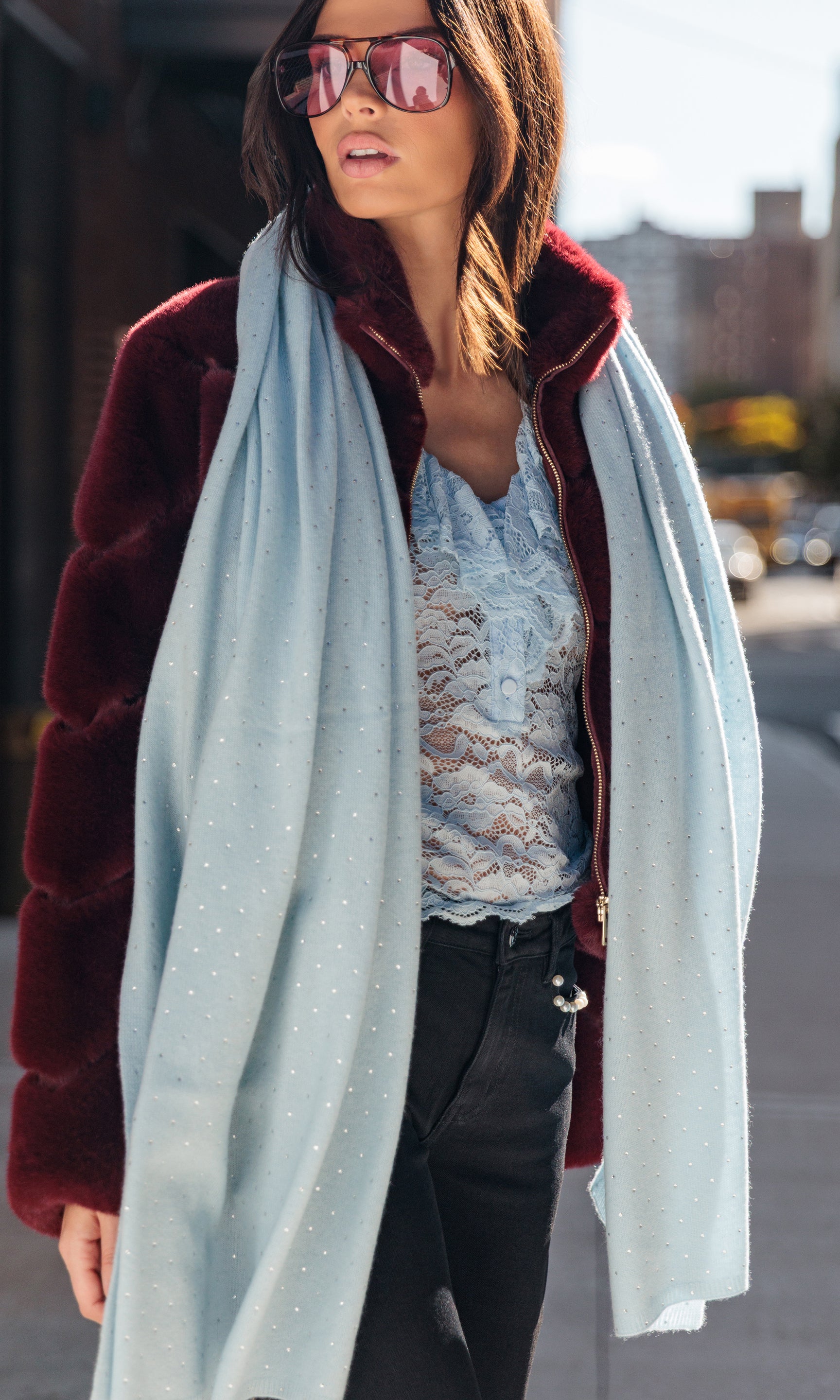 Woman wearing a burgundy coat with fur trim, light blue scarf, and sunglasses on a city street.