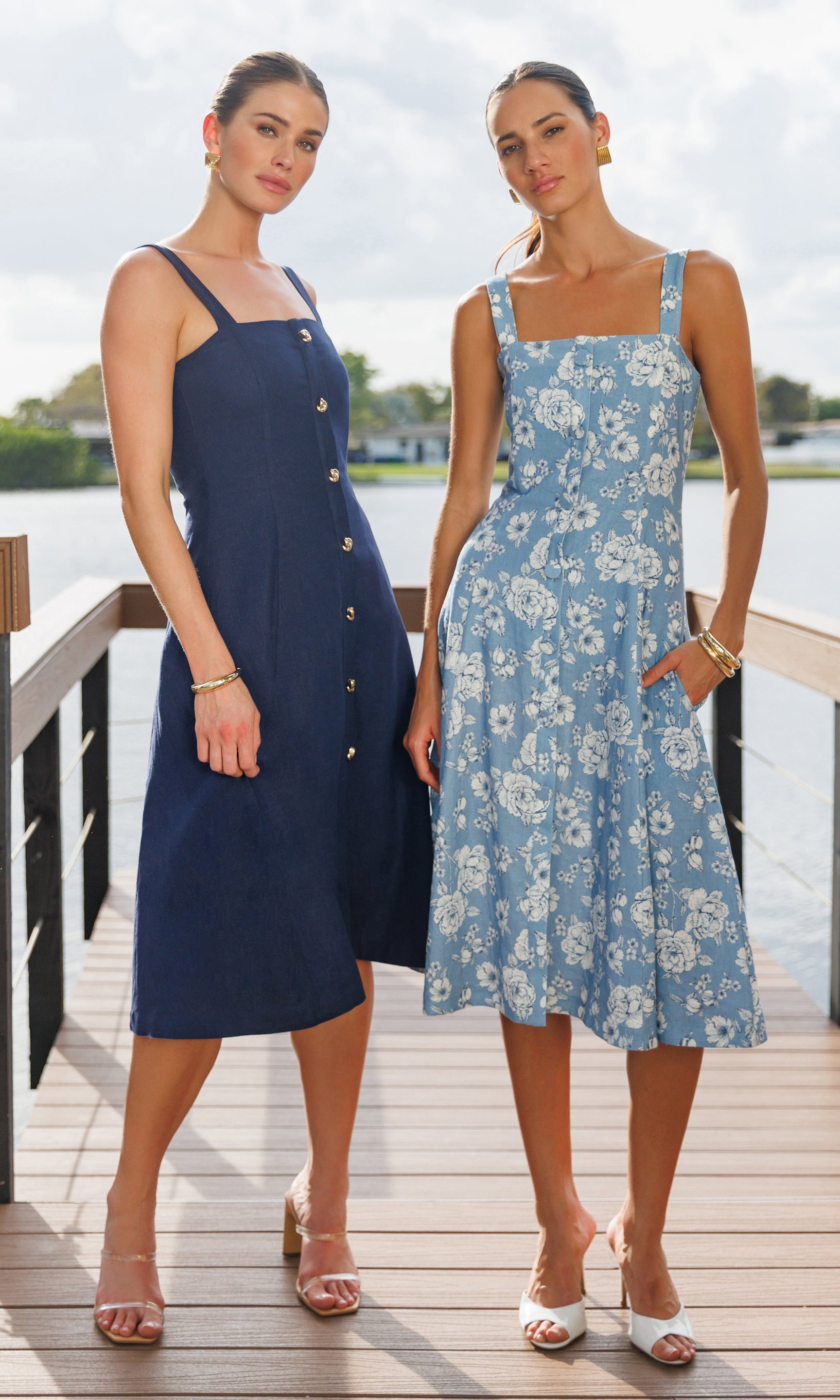 Two women wearing dresses standing on a dock by a body of water.