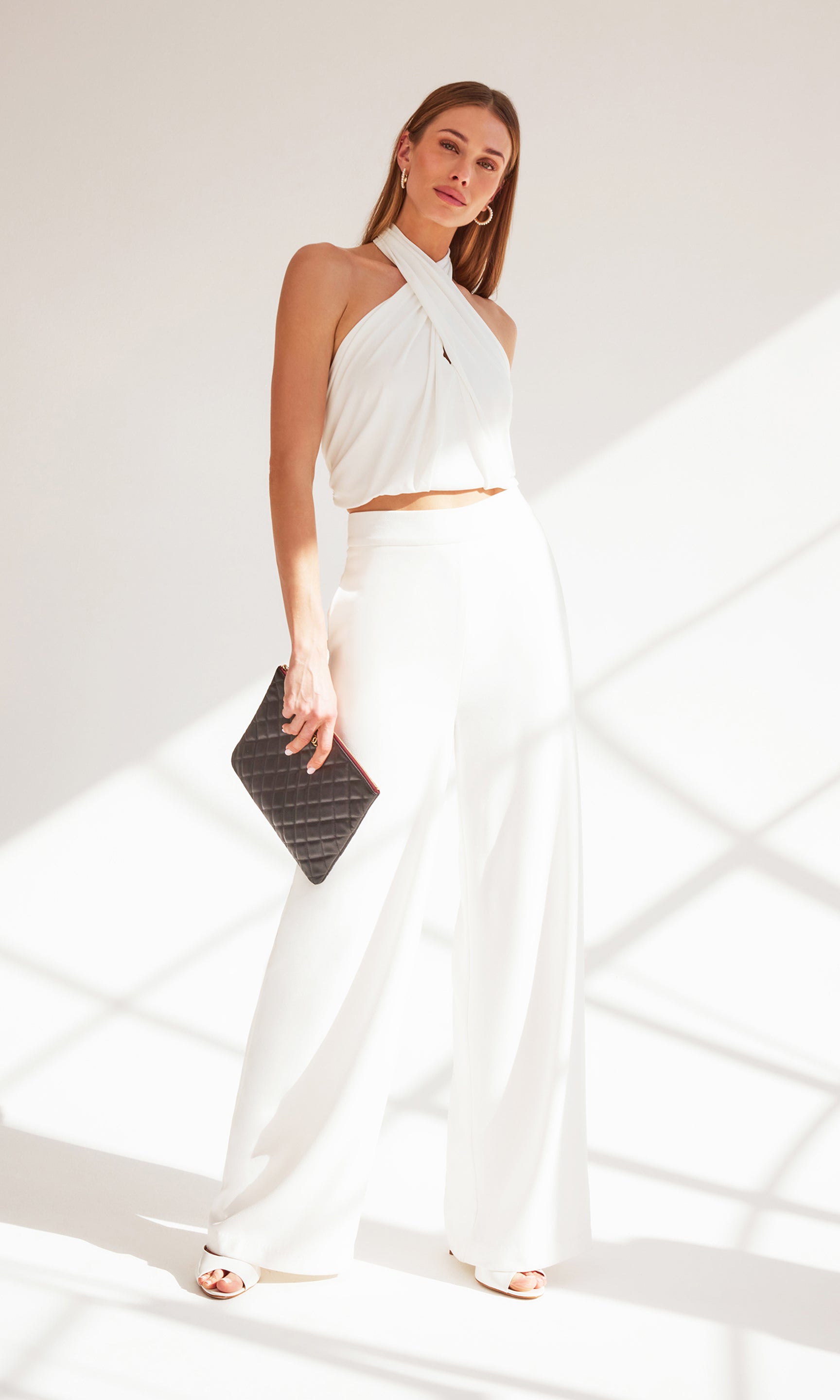 model posing in front of a window, wearing a white halter blouse paired with the white Magda trouser pants and white heels, holding a black clutch purse